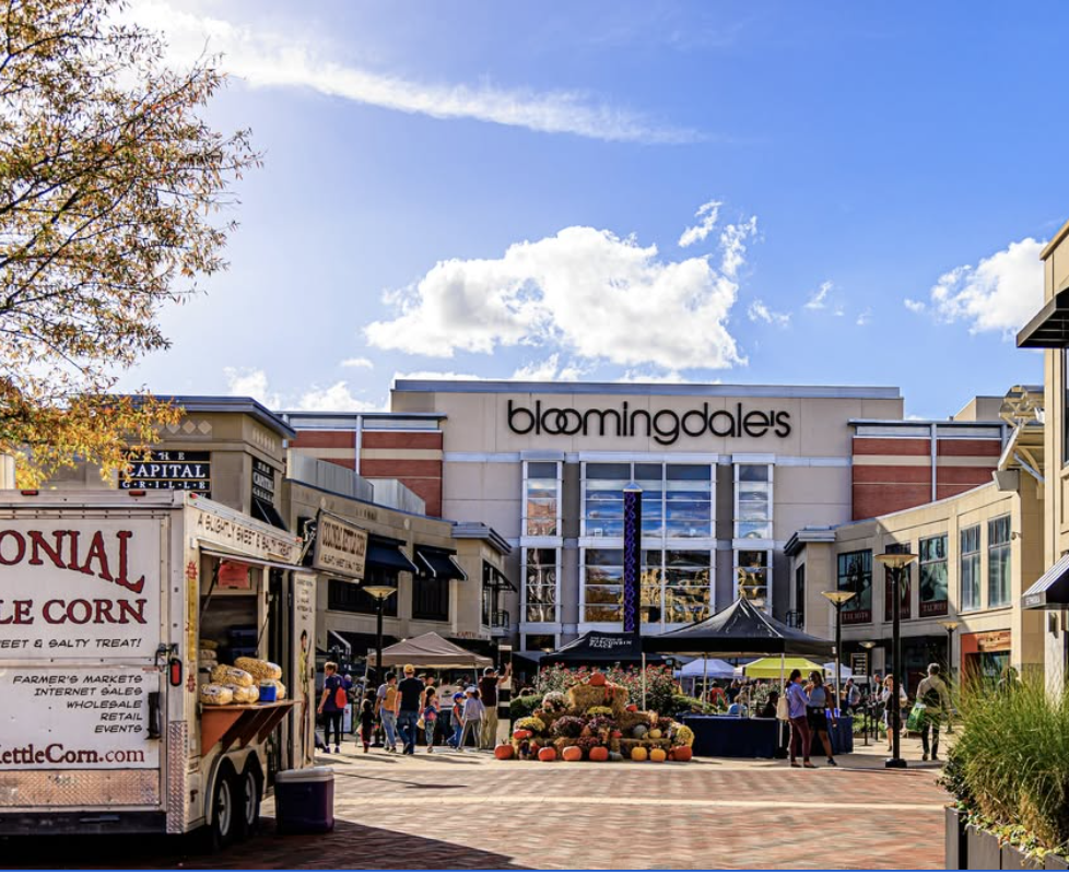 Bloomingdale's store with outdoor market stalls and shoppers in a shopping plaza on a sunny day.