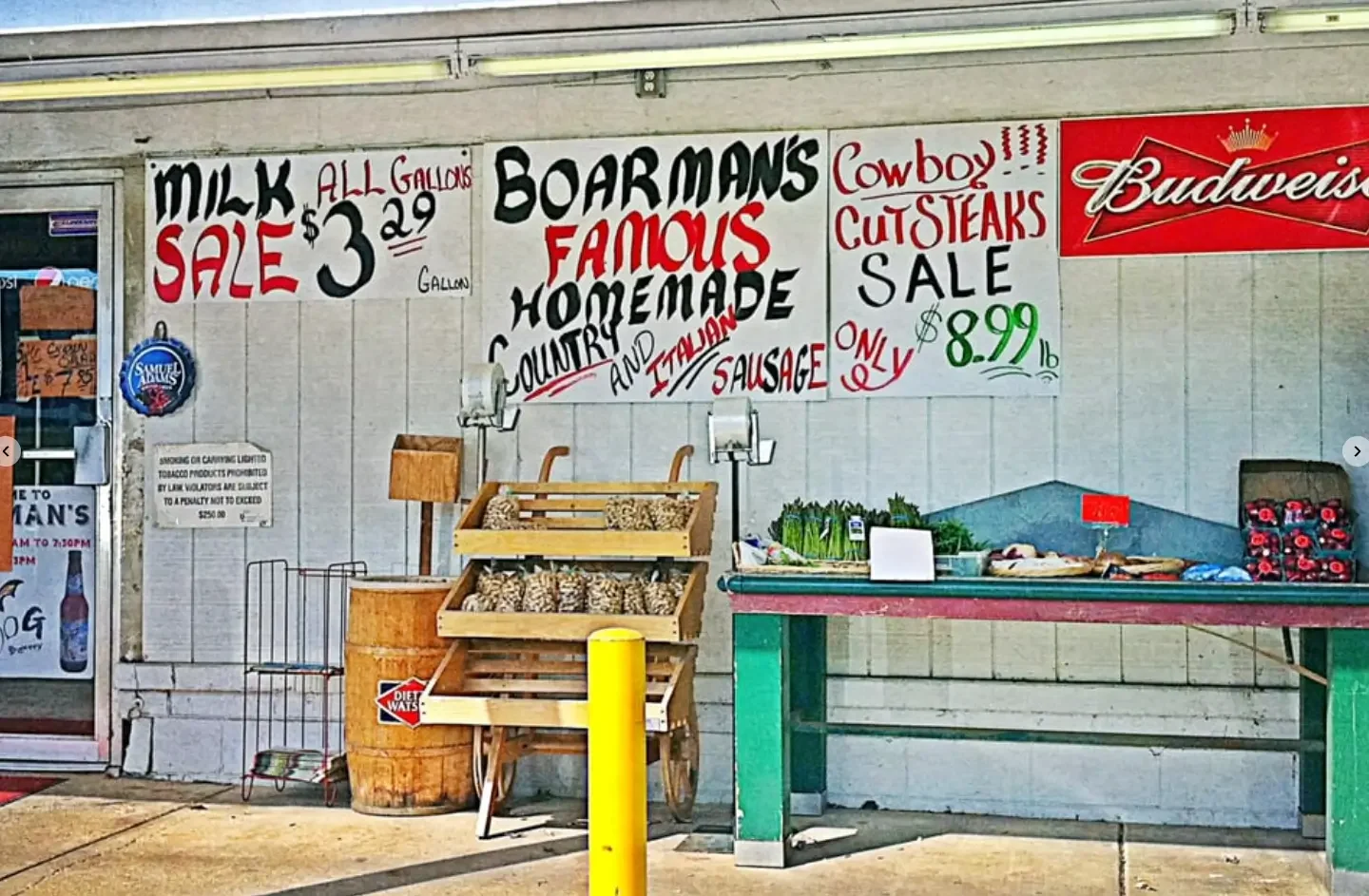 Outside of a small food stand with signs advertising a milk sale, homemade sausage, and a sale on cowboy cut steaks for $8.99. There is a table with vegetables and other food items, and a large wooden container with peanuts or similar snack, with a small cart nearby. A Budweiser sign and other promotional posters are visible.