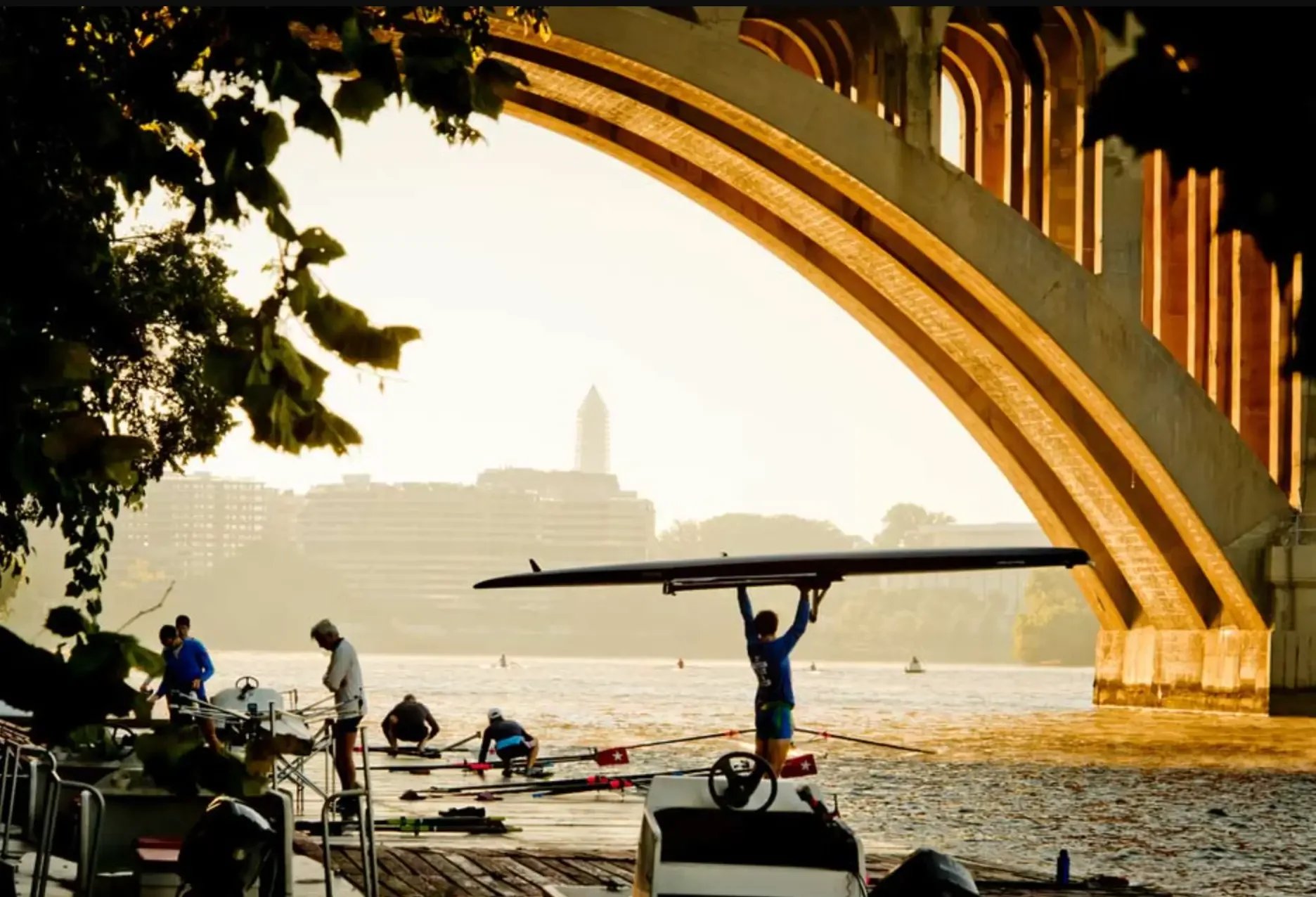 People preparing sailboats on a dock under an arched bridge over a river during sunset, with city buildings in the background.