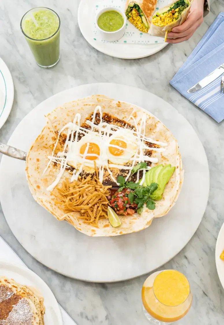 A plate with a tostada topped with two sunny-side-up eggs, shredded beef, avocado slices, pico de gallo, fresh cilantro, and a lime wedge, with a drizzle of white sauce.