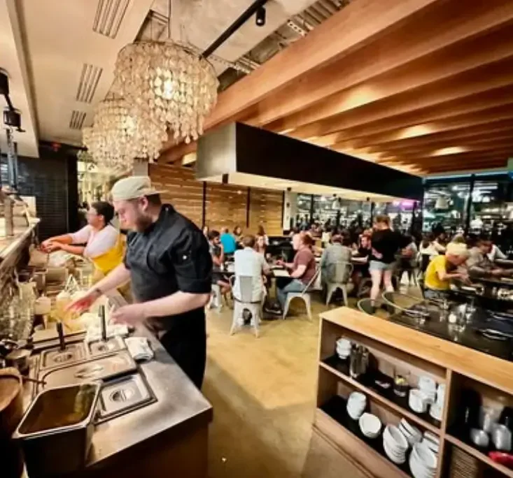 Busy restaurant with chefs preparing food at the counter and diners sitting at tables inside, featuring modern interior design with wood paneling and chandeliers.