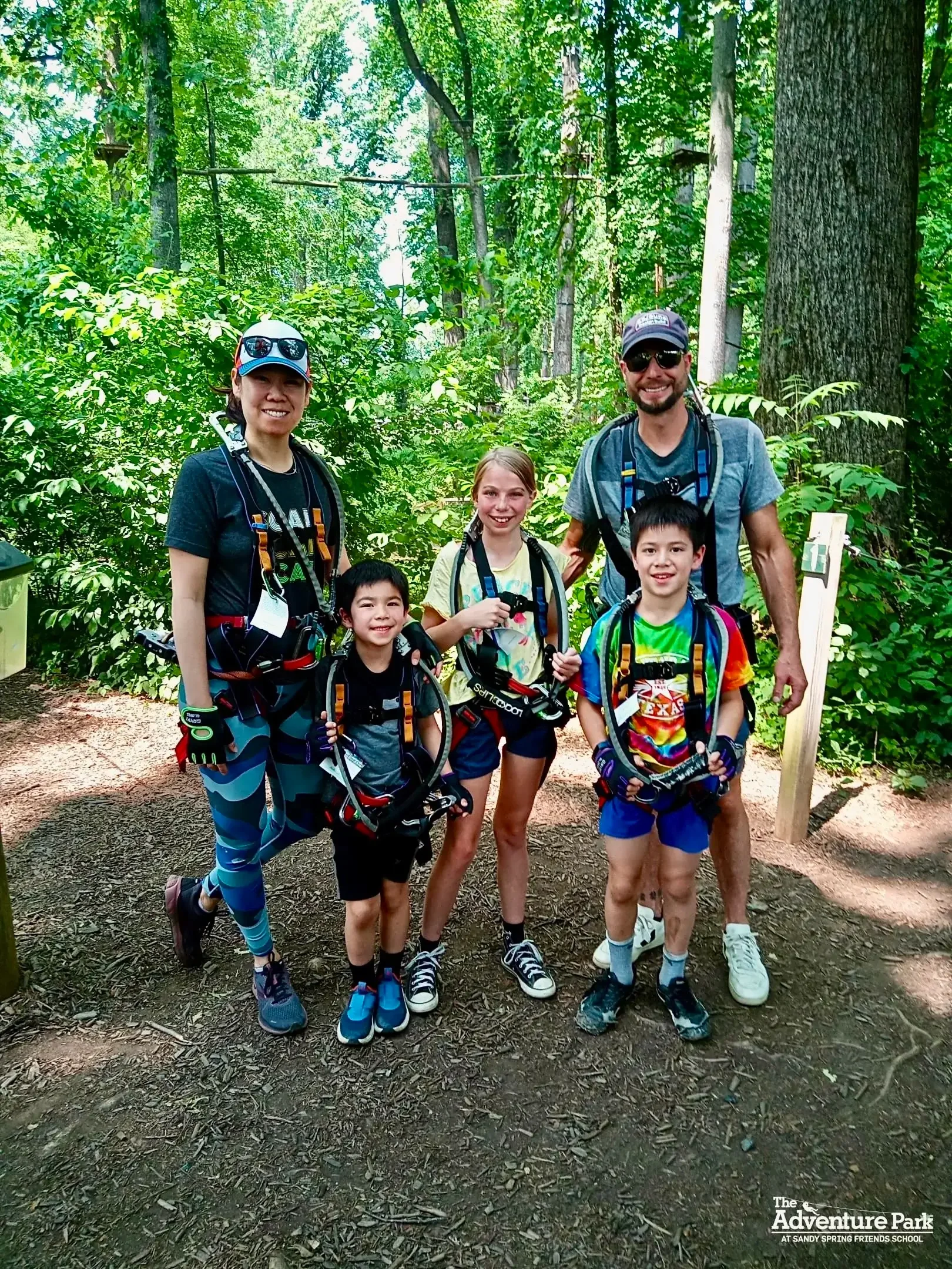A group of five people, two adults and three children, smiling and posing in a lush green forest during a zip-lining adventure. They are wearing harnesses and safety gear, ready for outdoor activity.