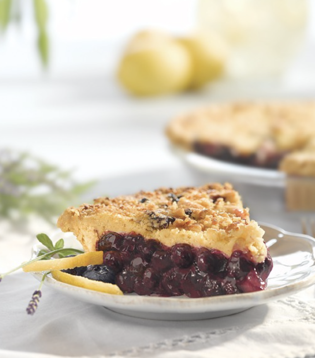 A slice of cherry pie on a white plate with lemon peel garnish, set on a textured white surface with blurred blueberries, lemon, and additional pie in background.