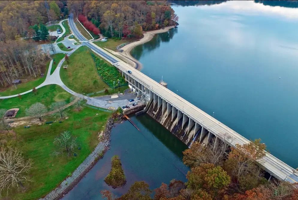 An aerial view of a dam and reservoir surrounded by green and autumn trees, with a winding road nearby and a park with walking paths.