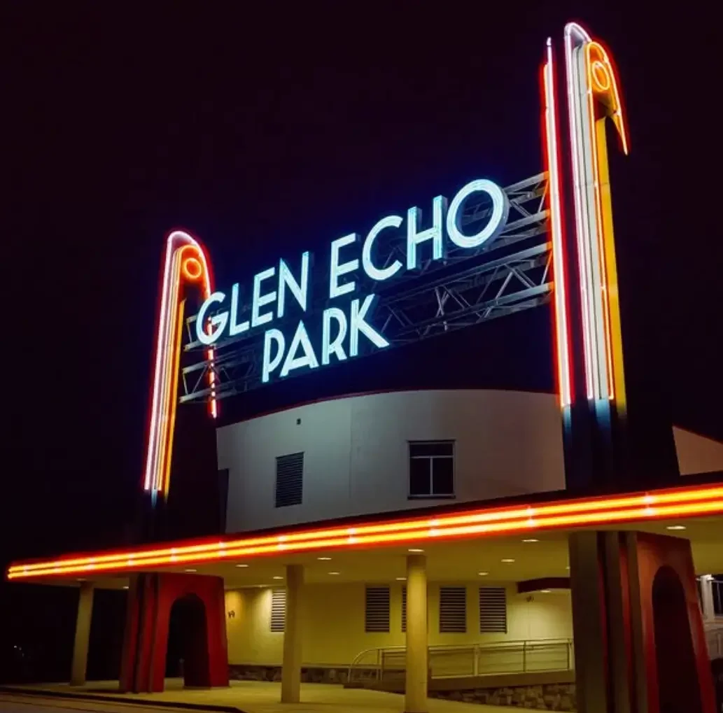 Neon sign reading "Glen Echo Park" at night, with vertical neon lights framing the sign, situated above a building entrance with columns and yellow walls.