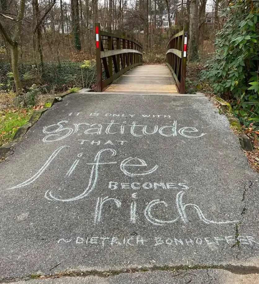 A paved pathway with a chalk message that reads, 'It is only with gratitude that life becomes rich,' leading to a small wooden footbridge over a creek, surrounded by trees and bushes.