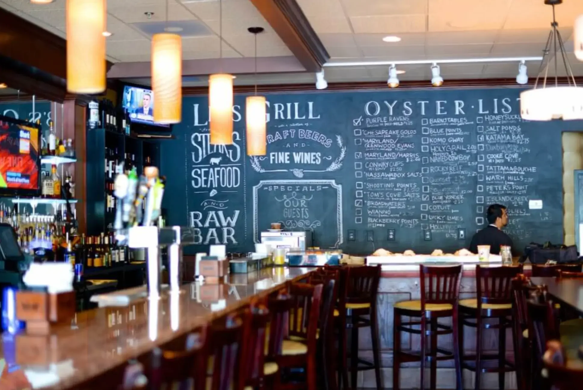 Interior of a seafood restaurant with bar seating, a chalkboard menu on the wall listing seafood and drinks, and a server attending to the bar area, illuminated by hanging lights.