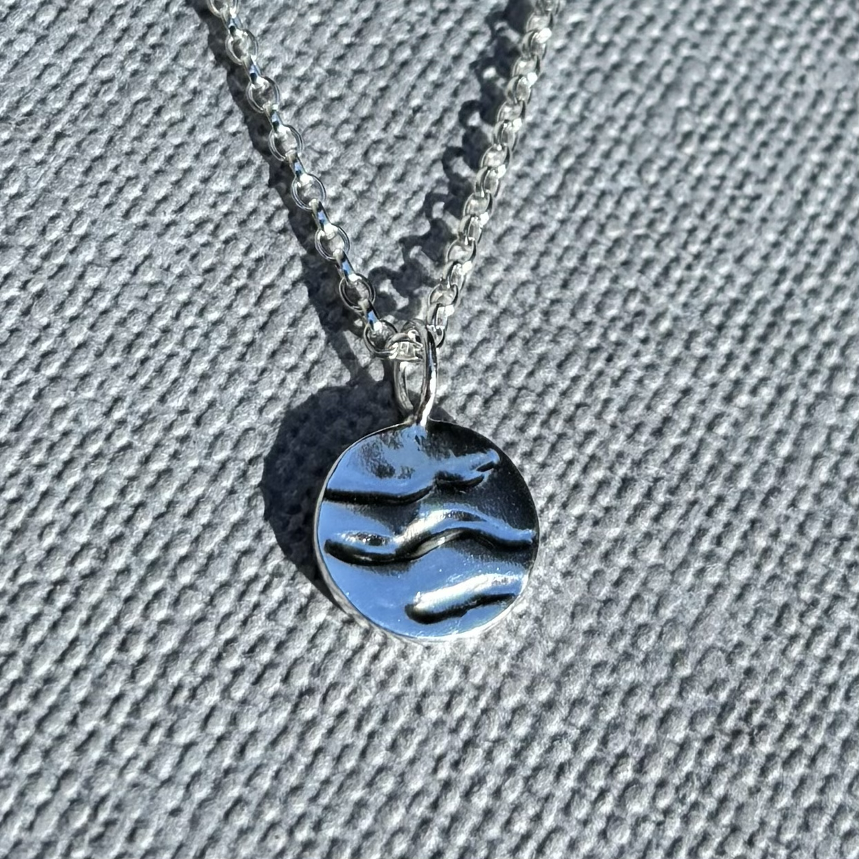 Silver necklace with a round pendant depicting 3 wavy grooves depicting waves lying on a textured gray surface.