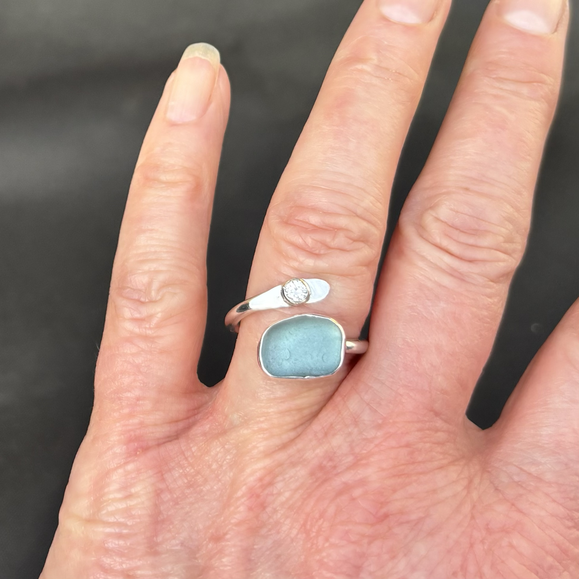 Close-up of a person's hand displaying multiple fingers with a silver open adjustable ring featuring a diamond and a pale blue piece of Dorset seaglass.