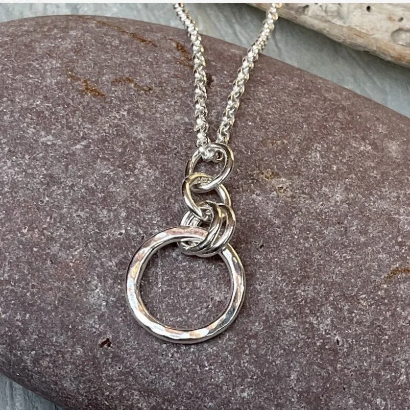 A silver necklace with several intertwined rings as the pendant, resting on a large rock with a branch in the background.