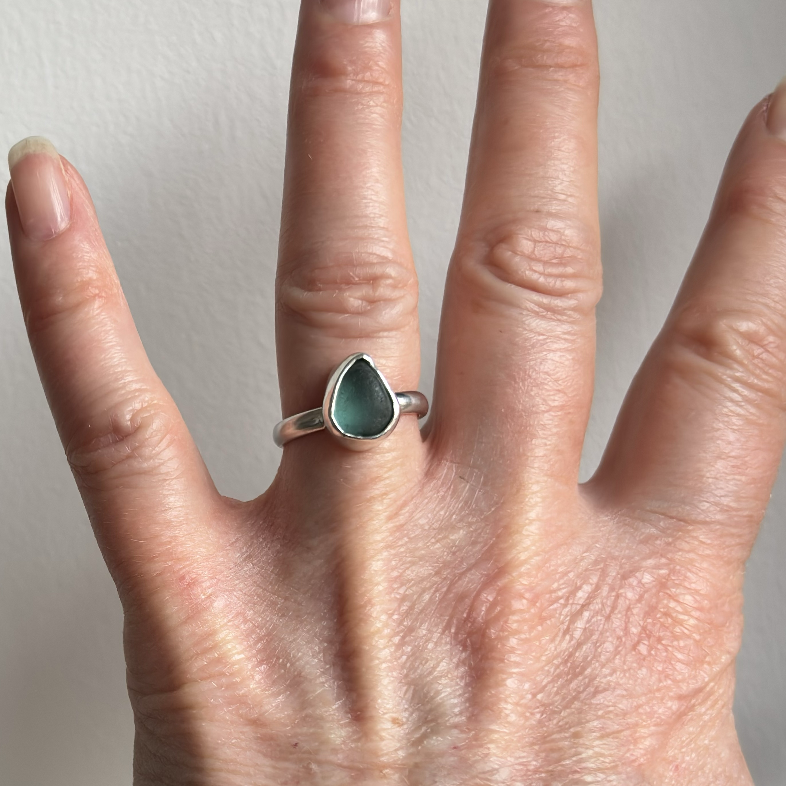 Close-up of a person's hand showing four fingers with one silver ring, featuring a blue pear shaped piece of seaglass, against a plain wall background.