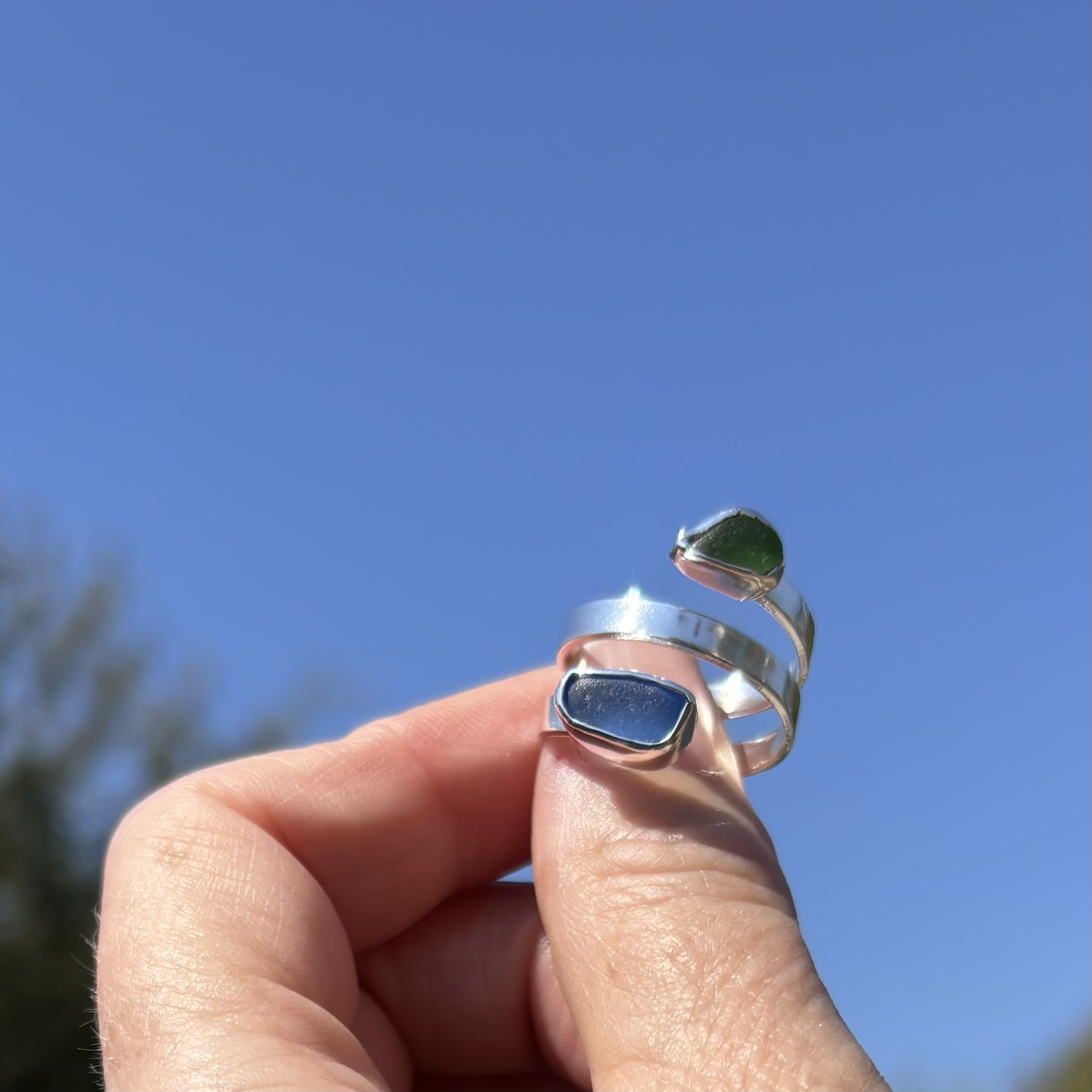 A hand holding a small shiny silver ring with blue and green seaglass against a clear blue sky with some blurred greenery in the background.