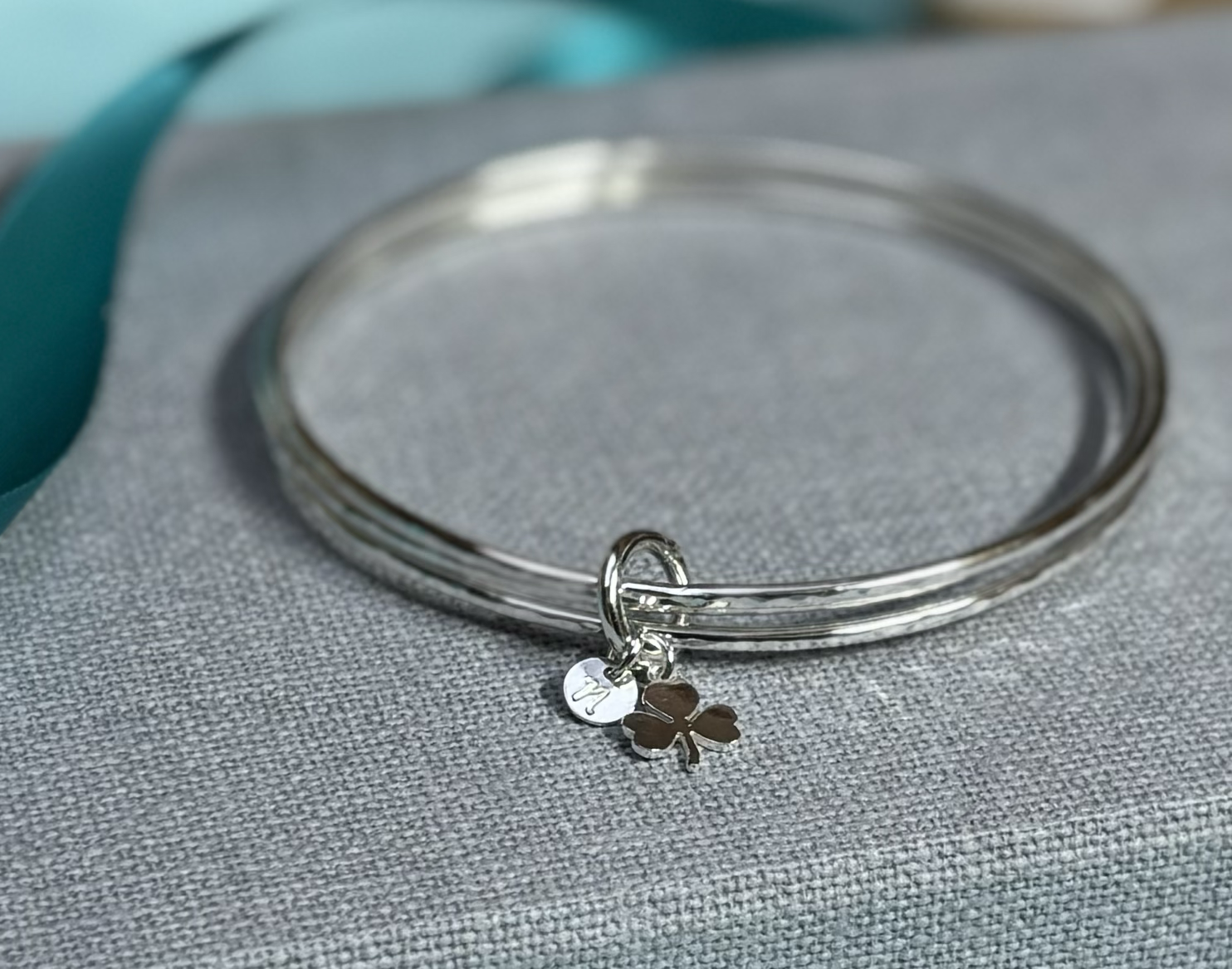 A silver bangle bracelet with small charms, including a shamrock and a circular charm with an engraved design, resting on a textured grey surface.