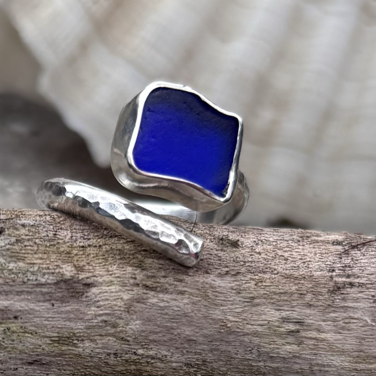 Close-up of an adjustable open silver ring with hammered textures, with a square-shaped deep blue pieceof Dorset seaglass, resting on a piece of weathered wood.