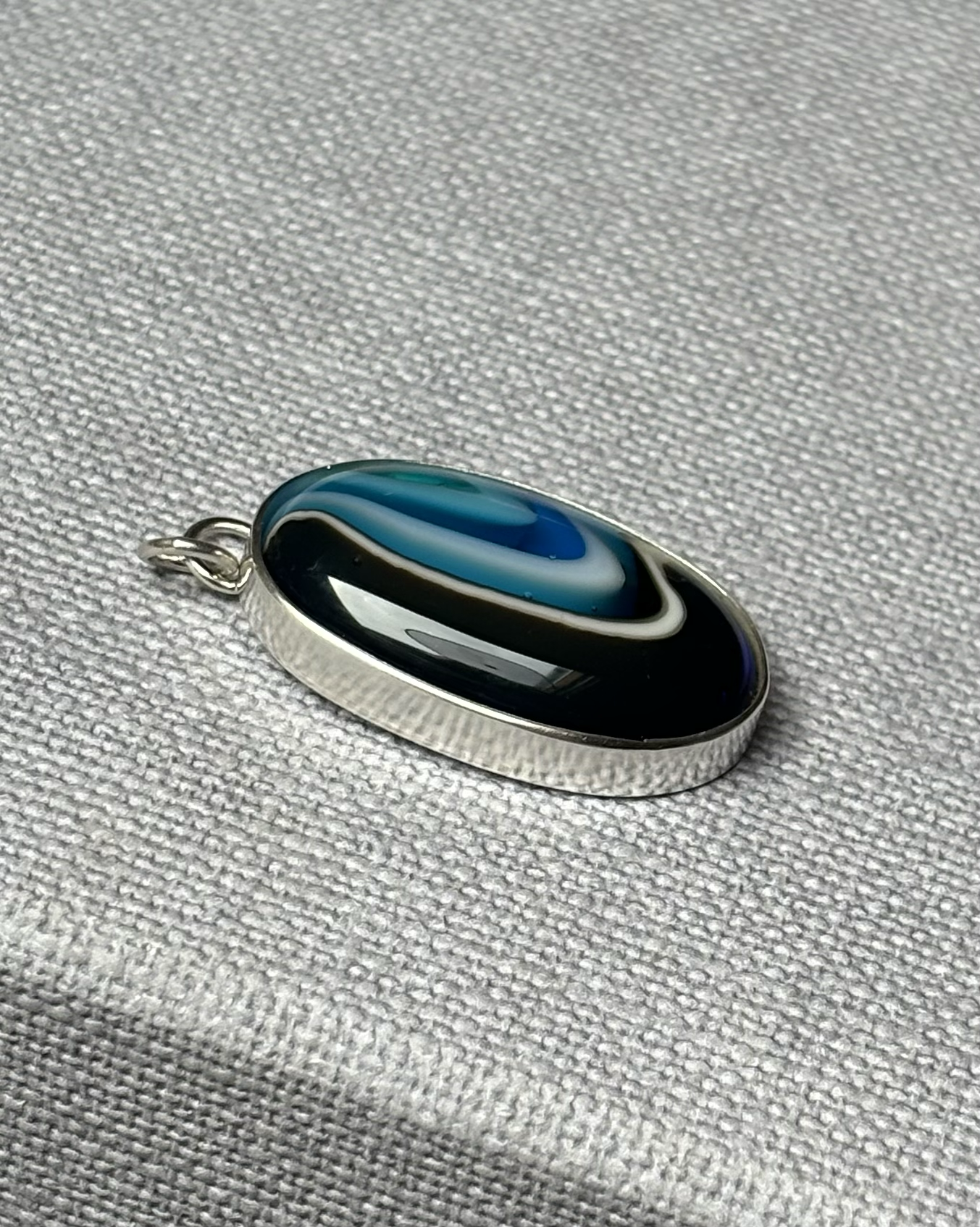 A silver pendant with a blue, white, and black glass marble design on a textured grey surface.