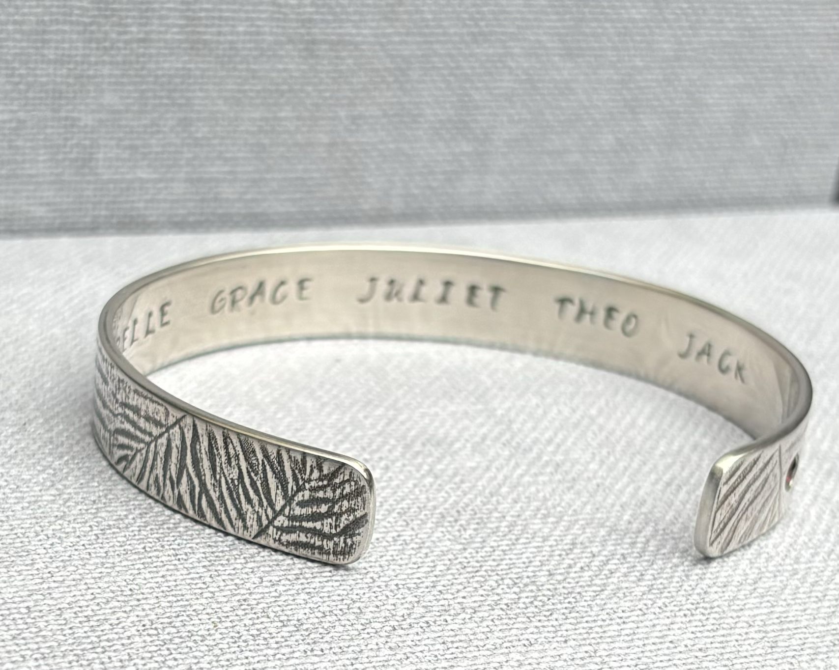 Sterling Silver cuff bracelet textured with a fern pattern and with engraved names on the inside, placed on a white textured surface.