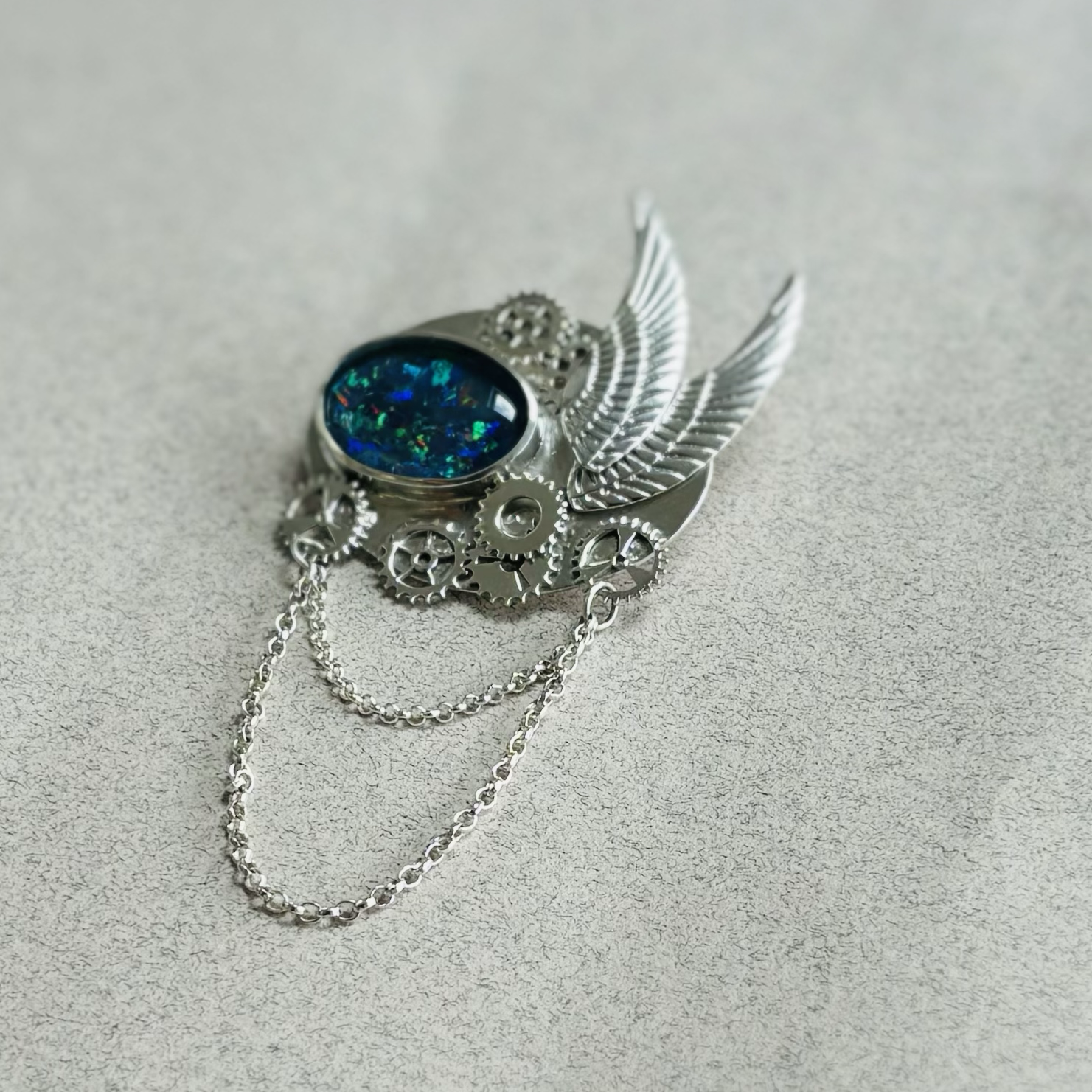 A steampunk-inspired brooch featuring a large blue opal gemstone set in a silver gear and wing design with chain details.
