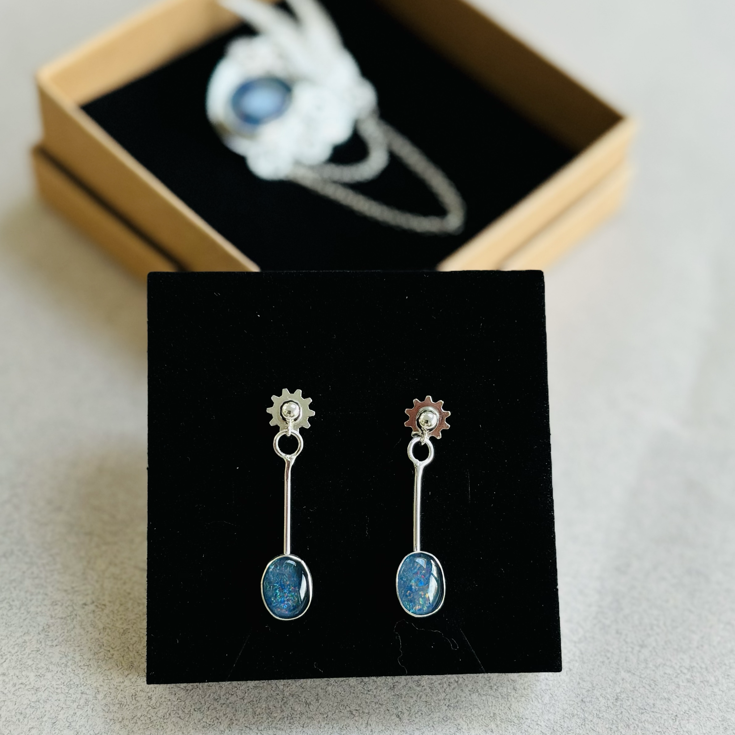 A pair of earrings with oval-shaped blue opal stones hanging from silver gear-shaped settings, displayed in a black jewellery box with a wooden lid.