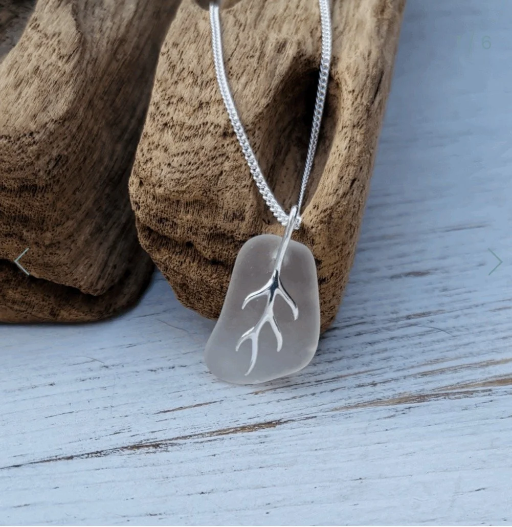 A Dorset seaglass necklace with sterling silver bail & detail shaped like a piece of seaweed or coral, hanging on a silver chain, displayed on a wooden surface.