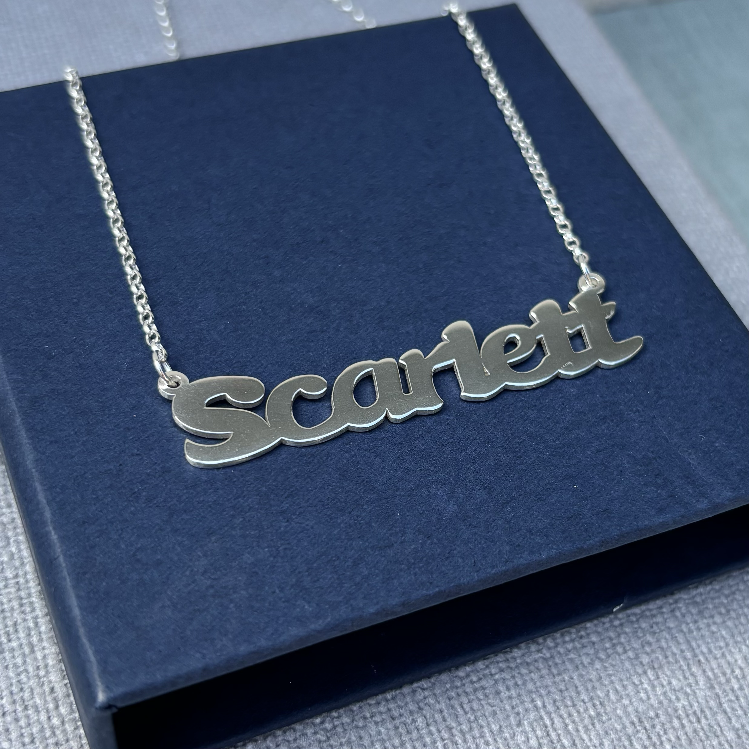 Close-up photo of a silver necklace with a pendant spelling the word Scarlett resting on a dark blue box.