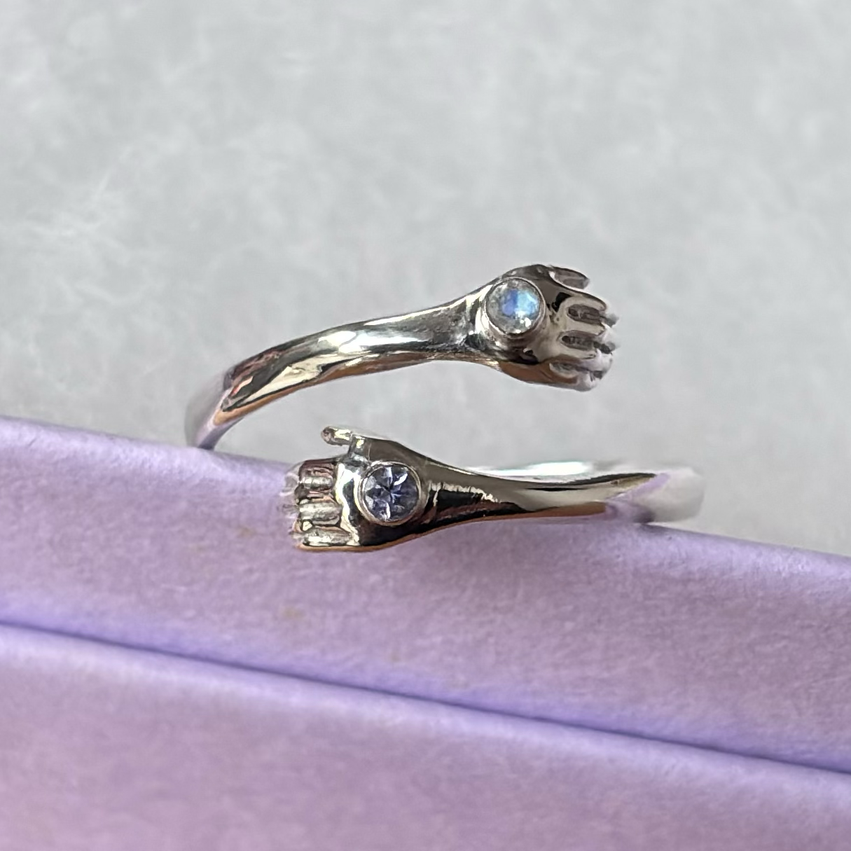 One silver open ring with small hand-shaped designs and embedded blue gemstones, resting on purple paper.
