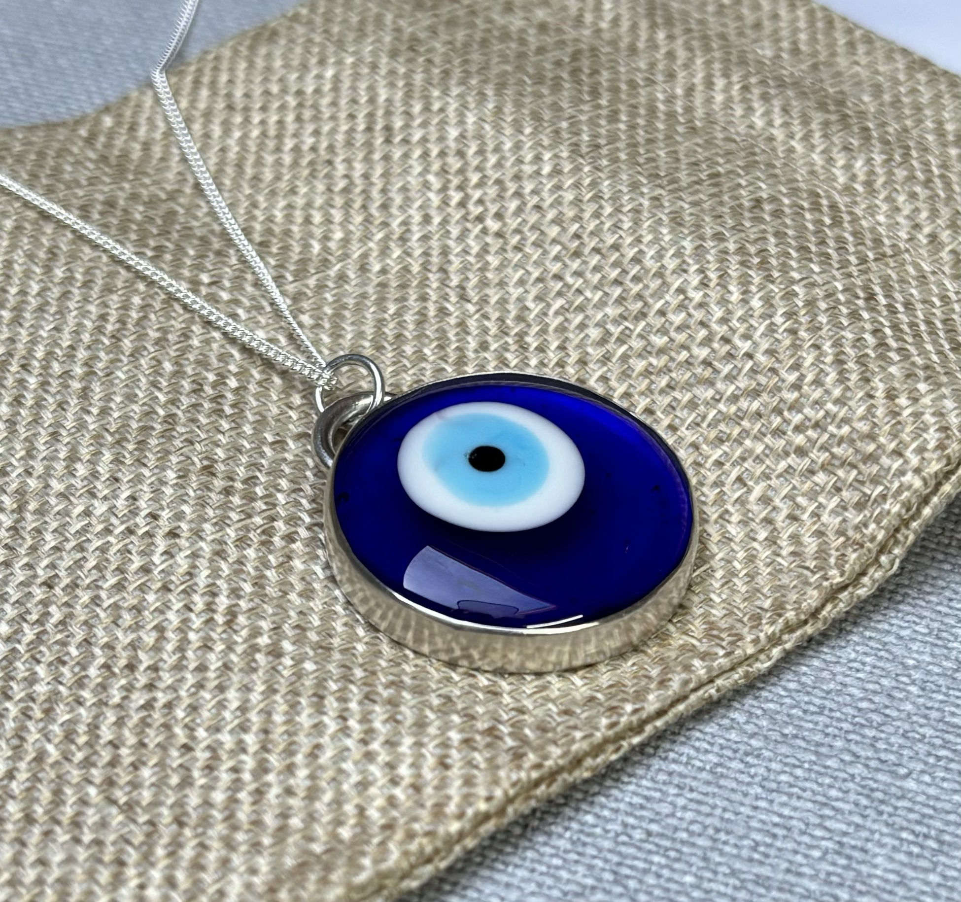 A close-up of an evil eye pendant on a silver chain resting on woven fabric. The pendant features a blue background with a white and black eye design in the centre.