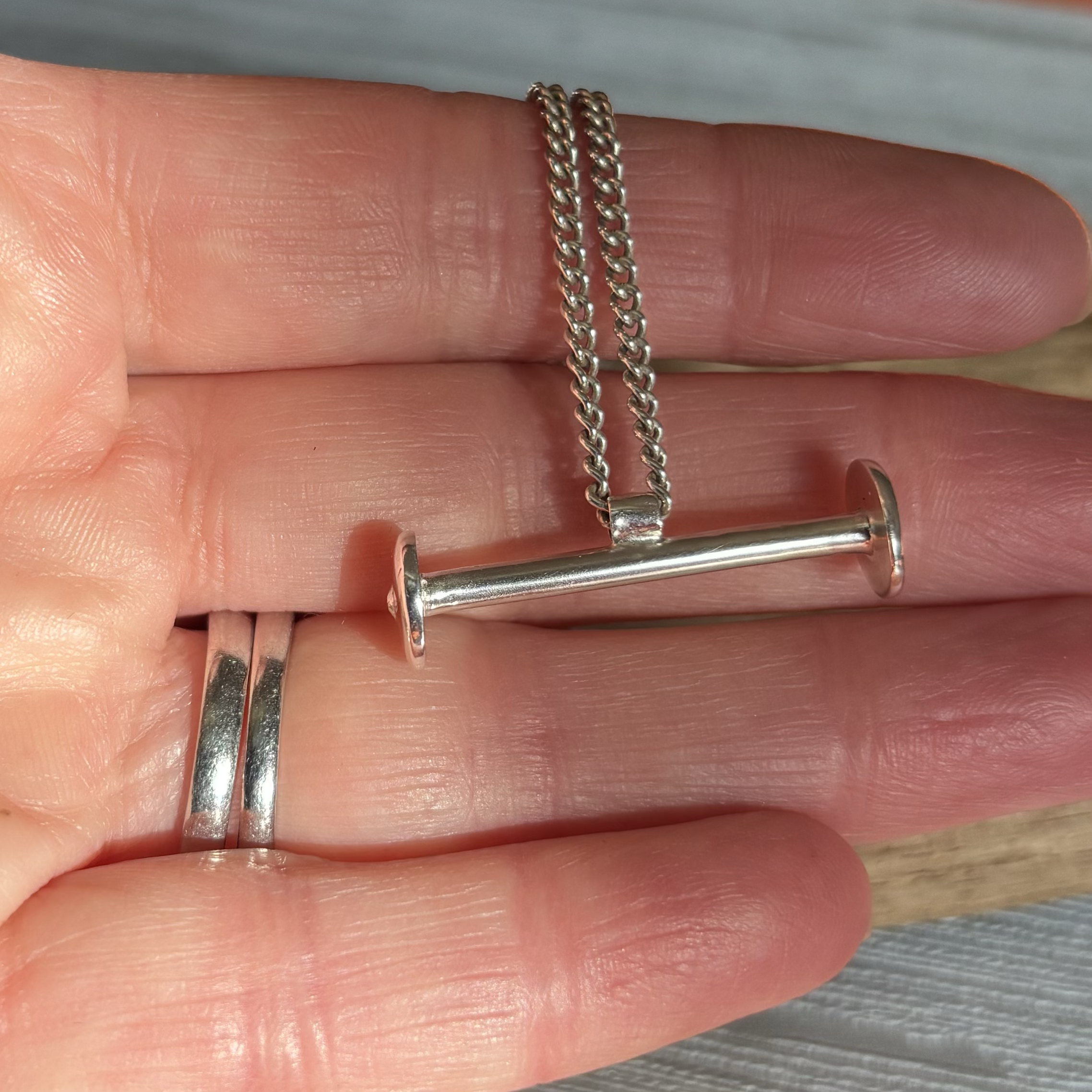 Silver chain pendant necklace in the shape of an old fashioned bobbin held between fingers.