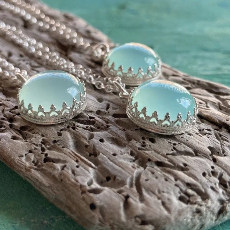 A pair of silver and turquoise coloured (Chalcedony) cabochon earrings with a decorative border of linked figures, resting on textured driftwood against a green background.