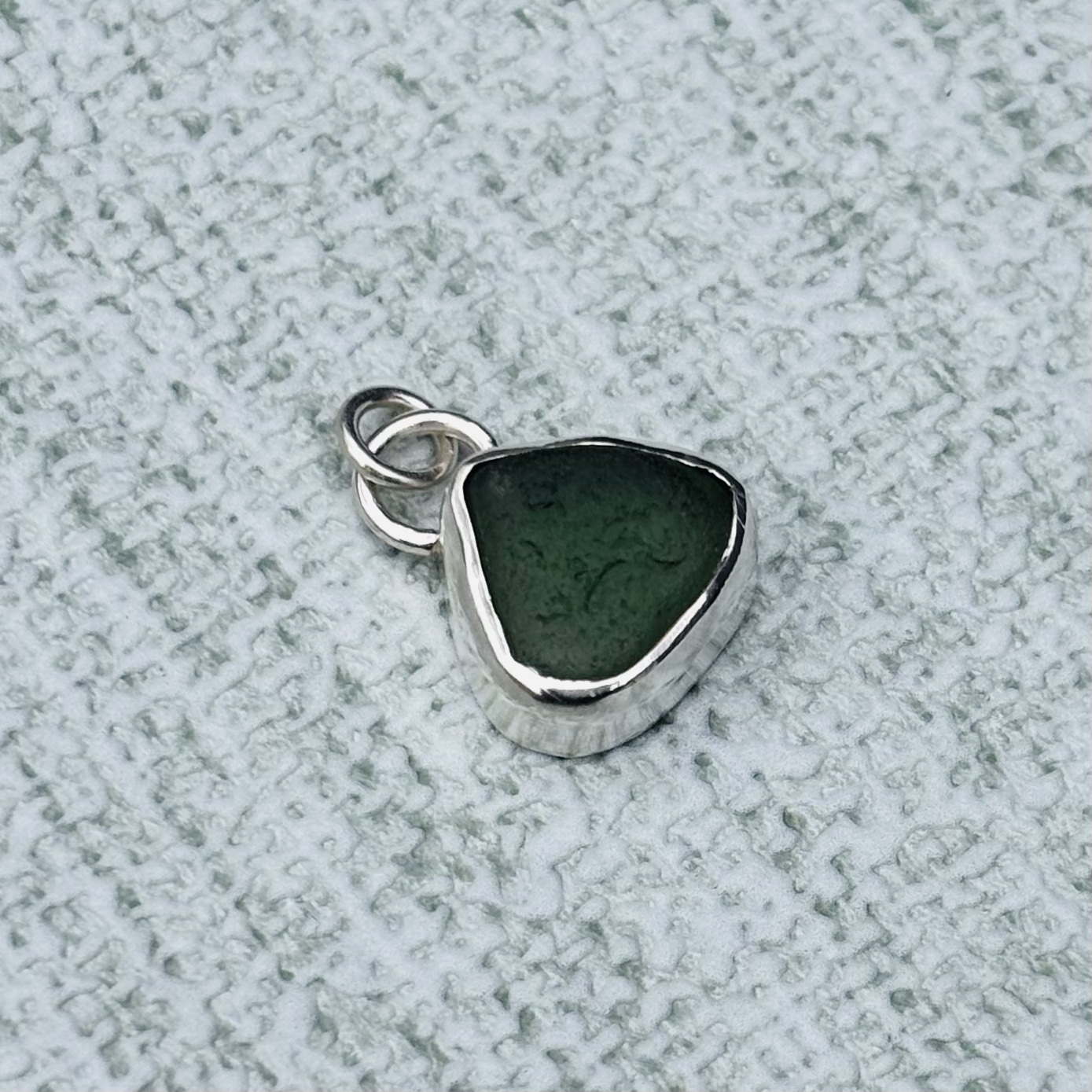A silver pendant with a dark green triangular seaglass stone on a textured grey surface.