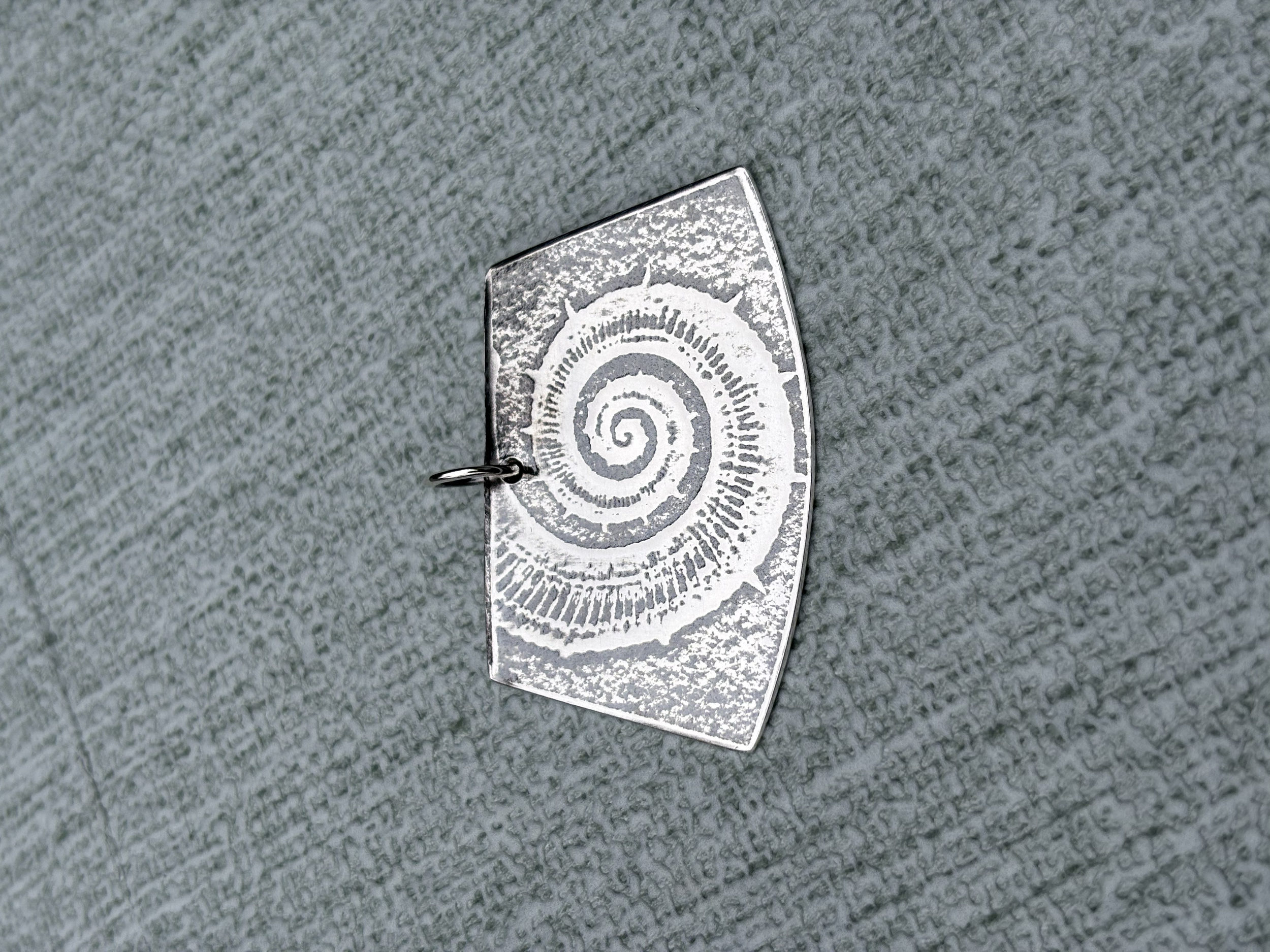 A silver pendant featuring an engraved Ammonite on it, placed on a textured grey surface.