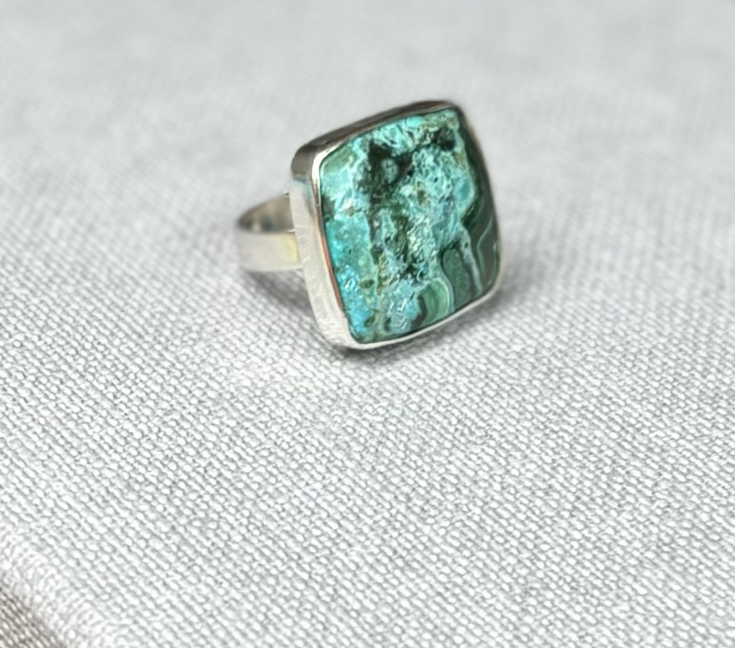 Silver ring with square turquoise stone on light grey fabric surface.