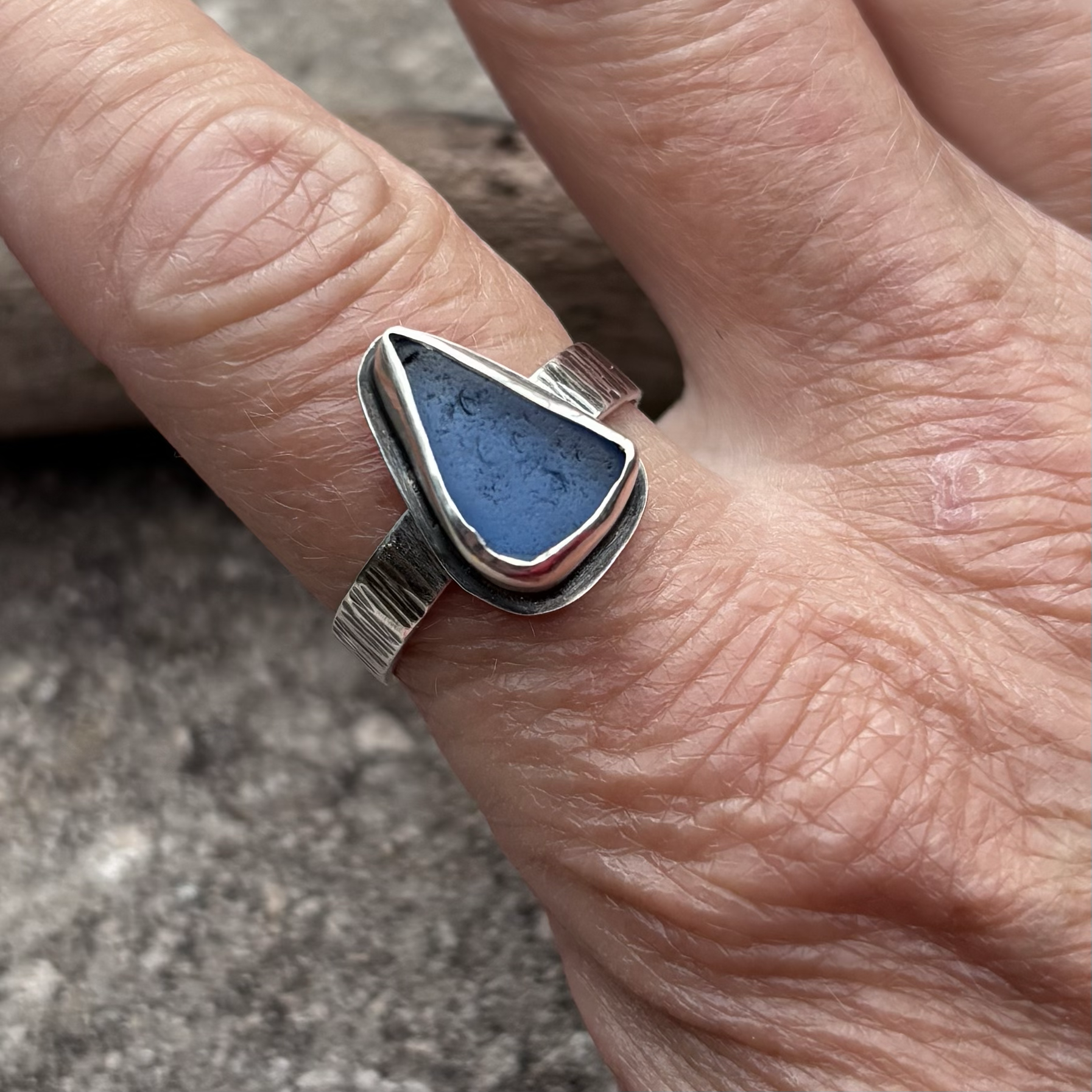 Close-up of a person's hand with a silver ring featuring a cornflower blue seaglass on the ring finger against a blurred background.