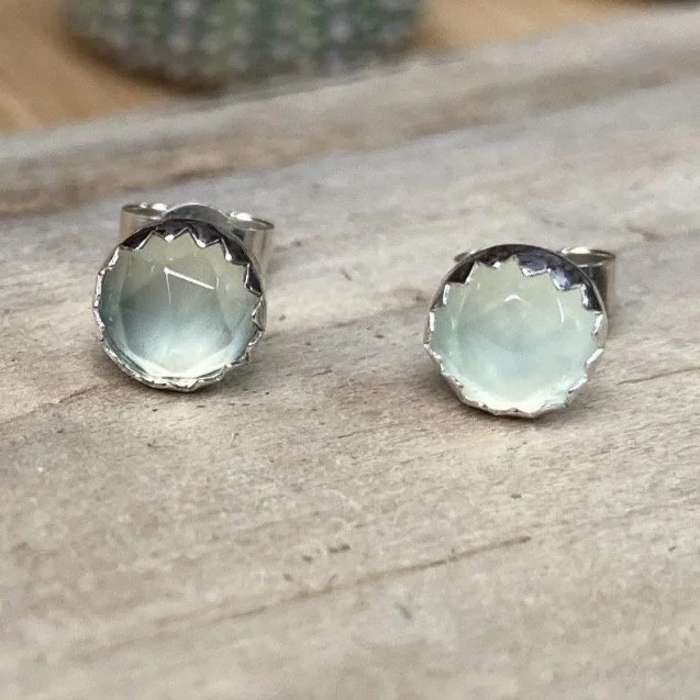Pair of silver earrings with round, pale greenish-white stone settings on a wooden surface.