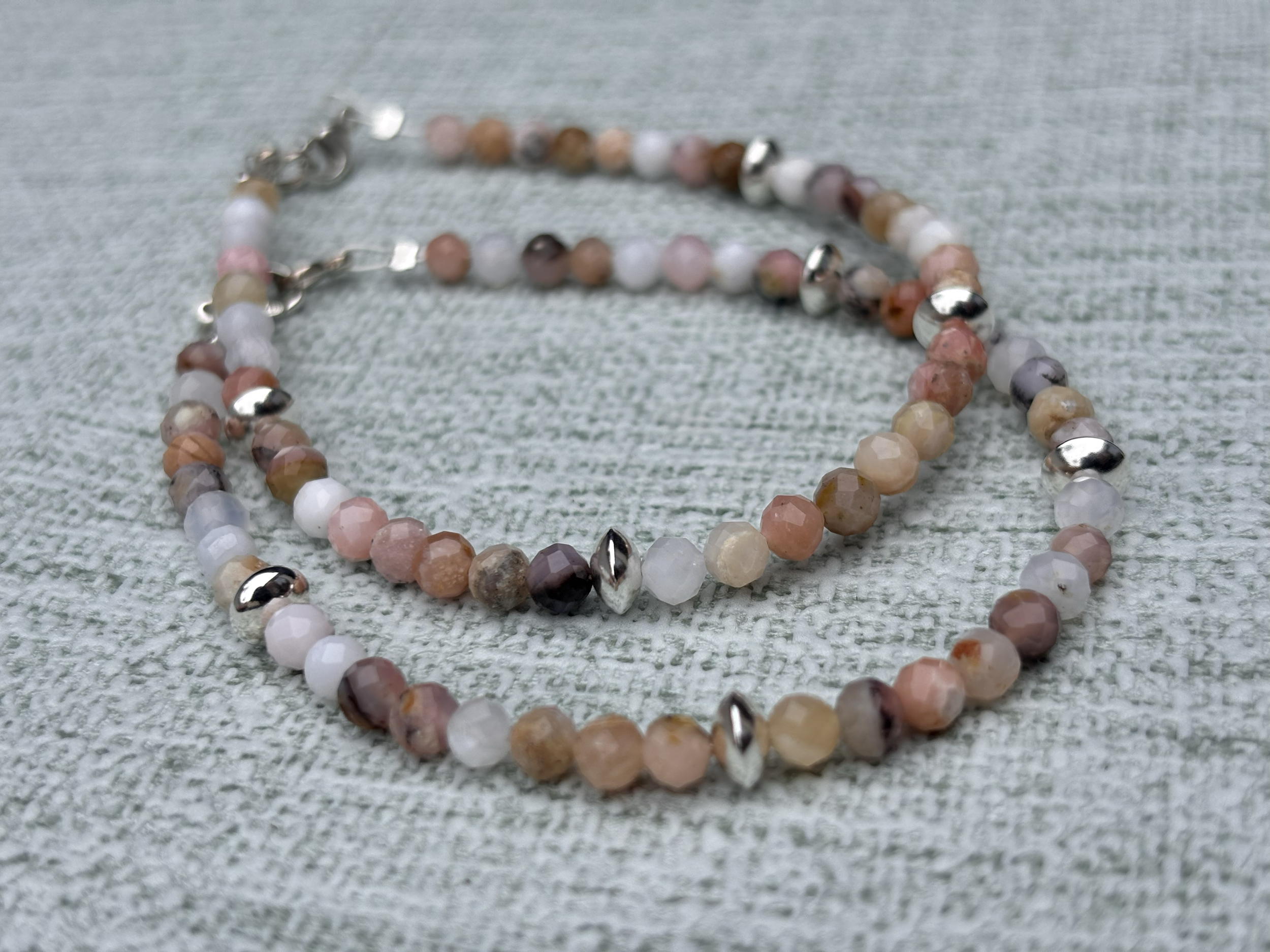 Close-up of a two Pink Opal beaded bracelets with multicoloured gemstone beads and silver accents, lying on a textured, light-coloured fabric surface.