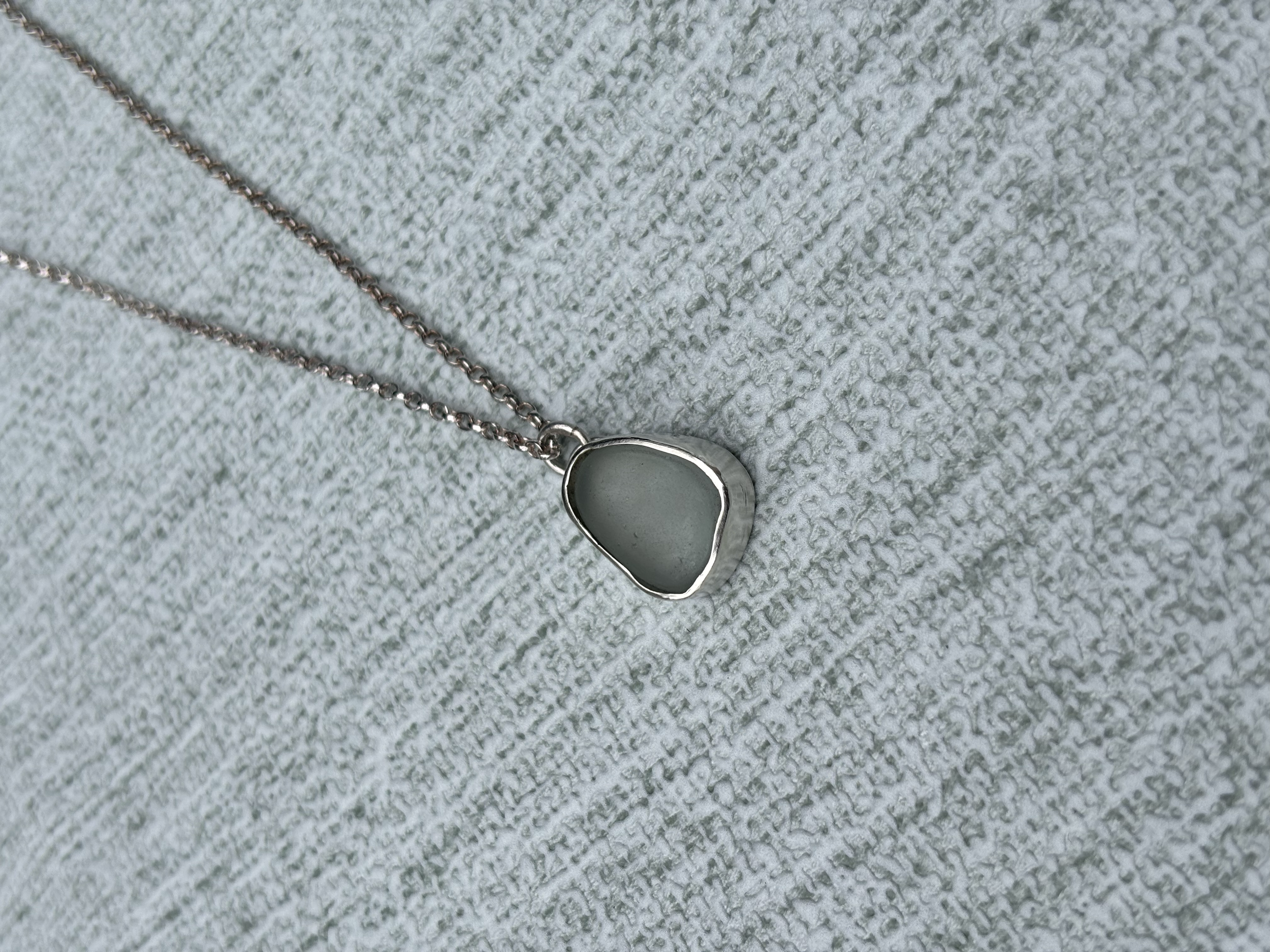 A silver necklace with a pale blue grey seaglass pendant lying on a textured, light-coloured fabric surface.