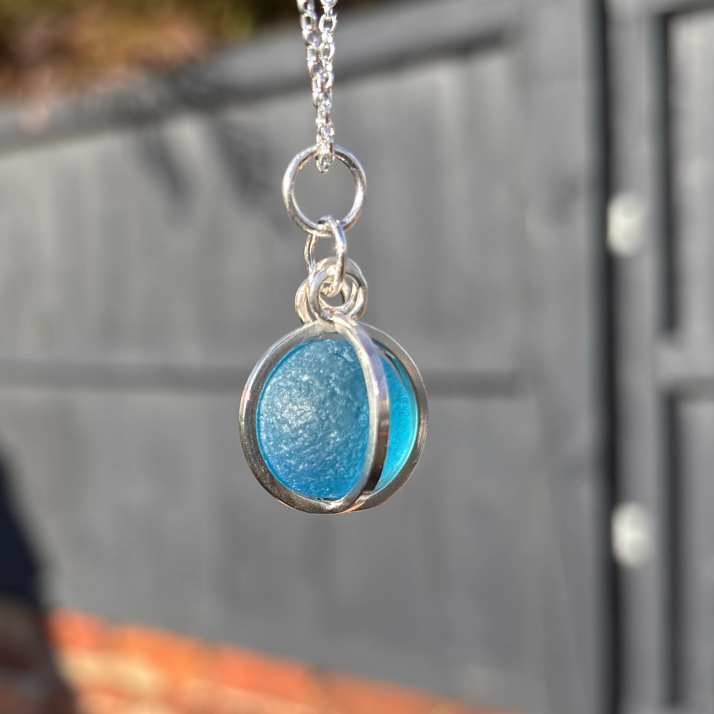 A close-up of a necklace with a round pendant containing blue seaglass marble, hanging against a blurred background.