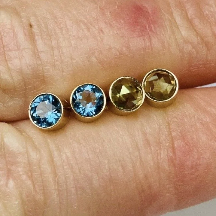 Four stud earrings on a finger, with two featuring blue gemstones (Topaz) and two featuring yellow-green gemstones (Peridot), all in gold settings.