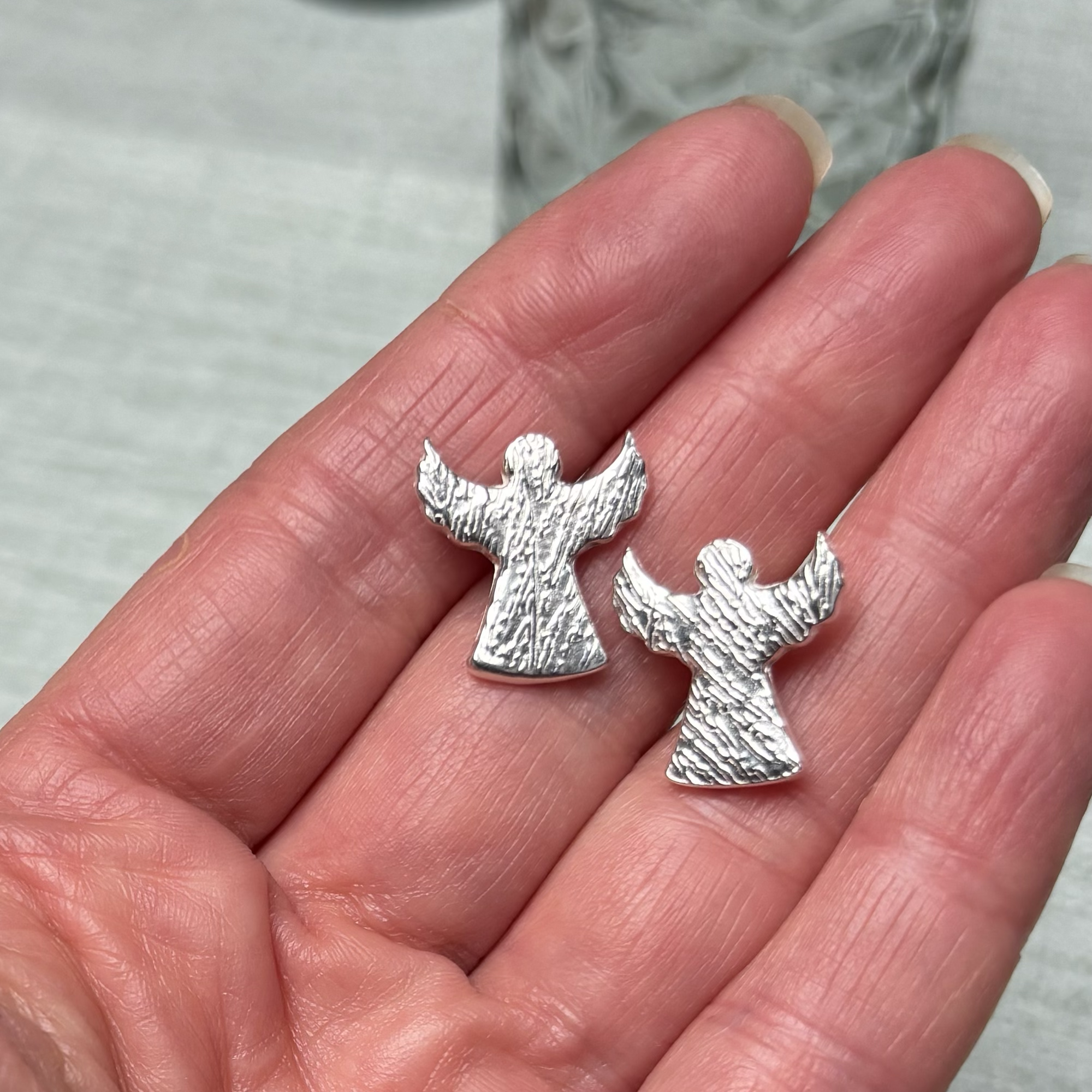 Close-up of a hand with a pair of angel shaped earrings made of feathered textured fine silver, set against a blurred background.