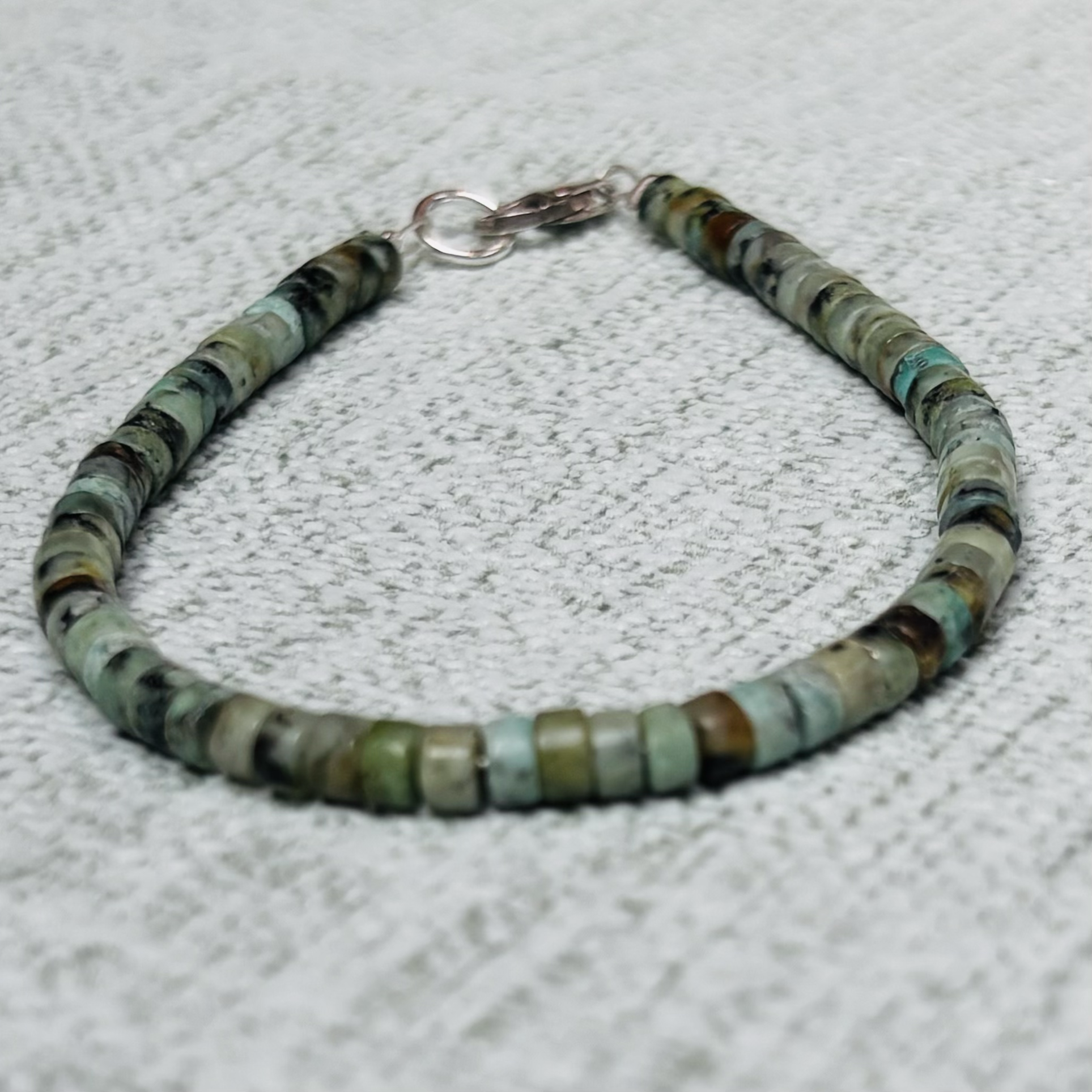 Close-up of a beaded necklace with round African Turquoise beads in shades of green, blue, grey, and brown, connected by a silver clasp, lying on a textured light grey surface.