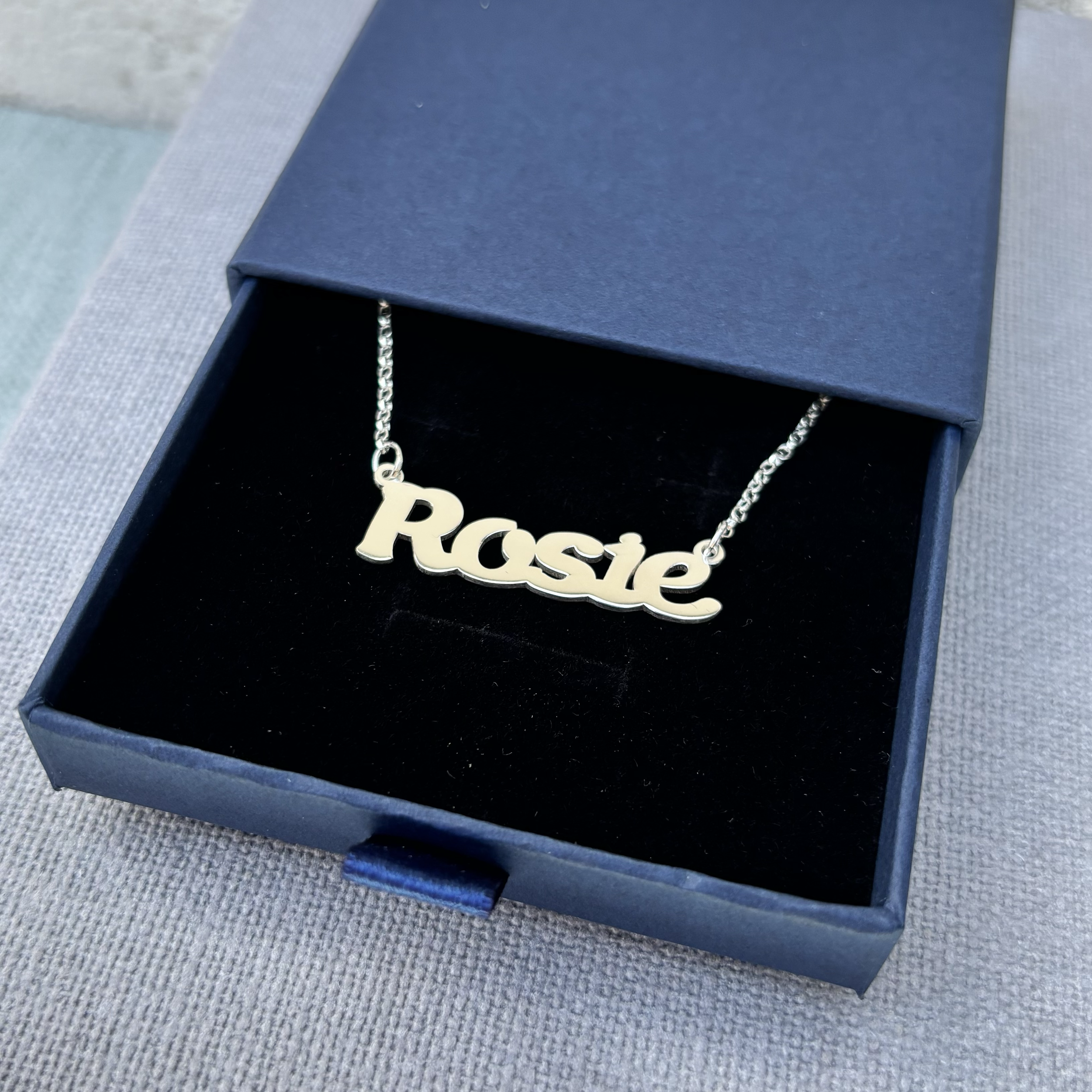 Close-up of a blue jewellery box with a silver chain necklace featuring a pendant that spells the word 'Rosie'.
