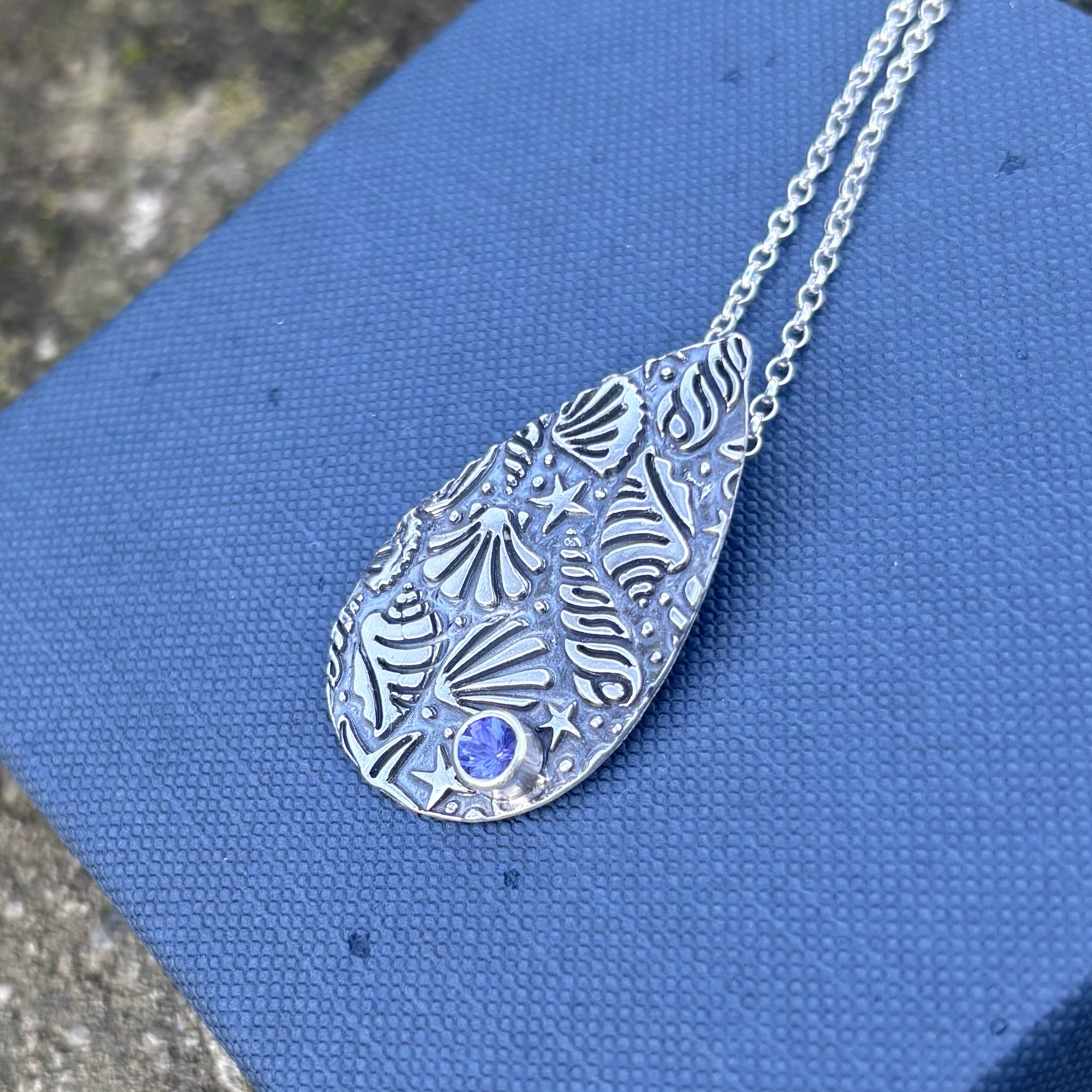 Silver necklace teardrop necklace with texture and a purple gemstone resting on a textured blue surface, with a blurred background.
