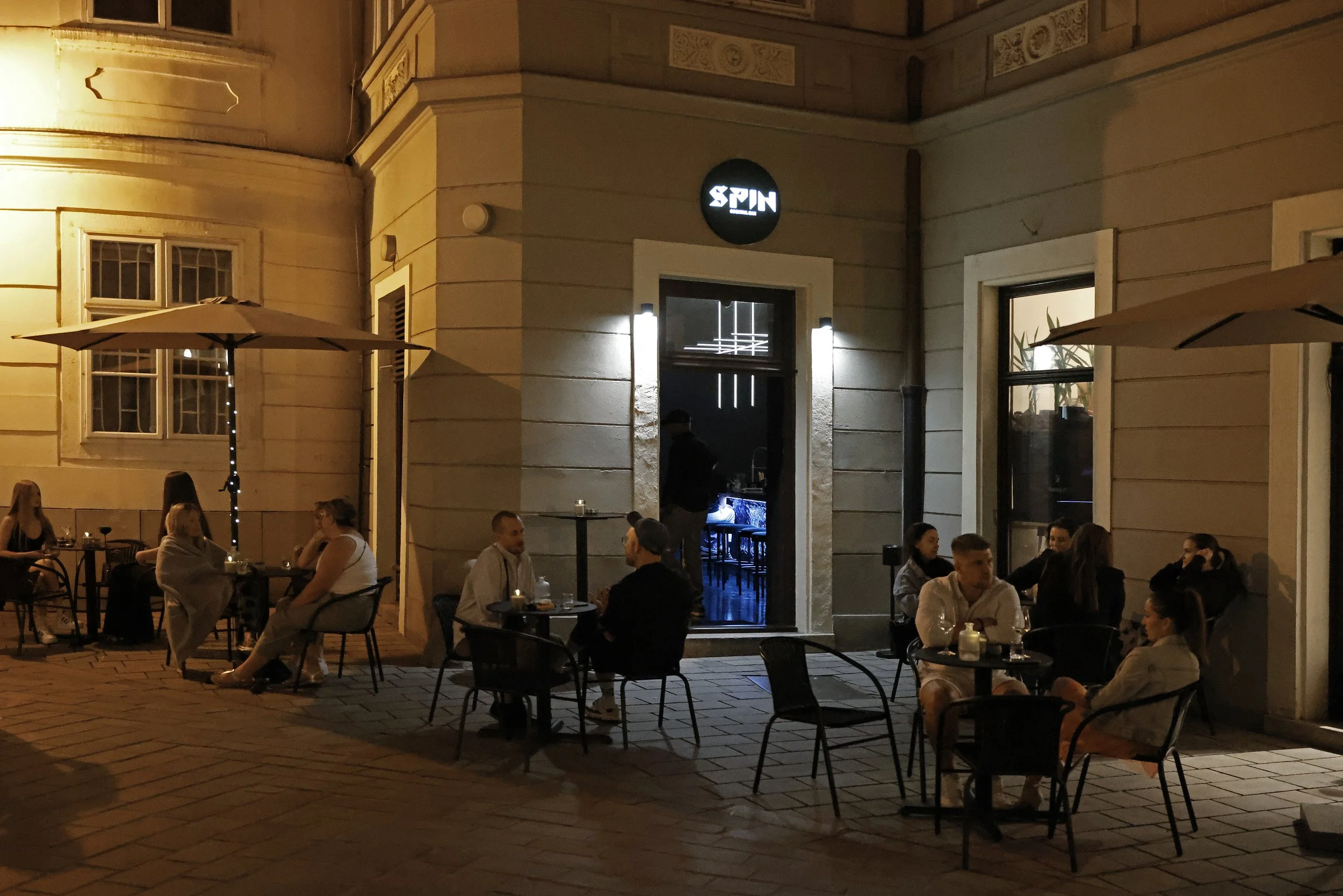 An outdoor patio at night with several groups of people sitting at tables, some with drinks, under umbrellas with a building in the background. There is a lit sign that says 'SPIN' above a doorway.