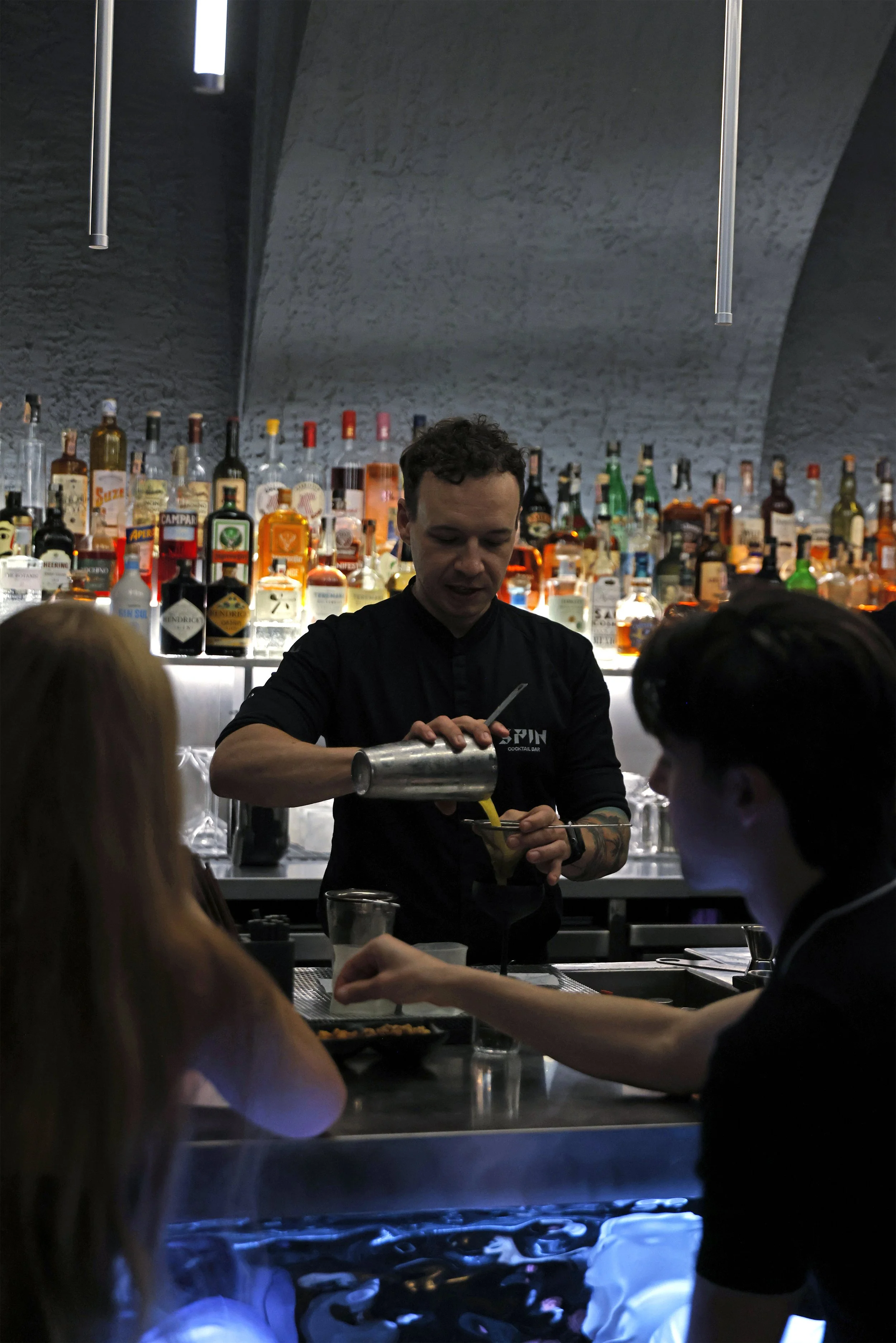 Bartender pouring an yellow alcoholic drink for two customers at a dimly lit bar with a backlit liquor display.