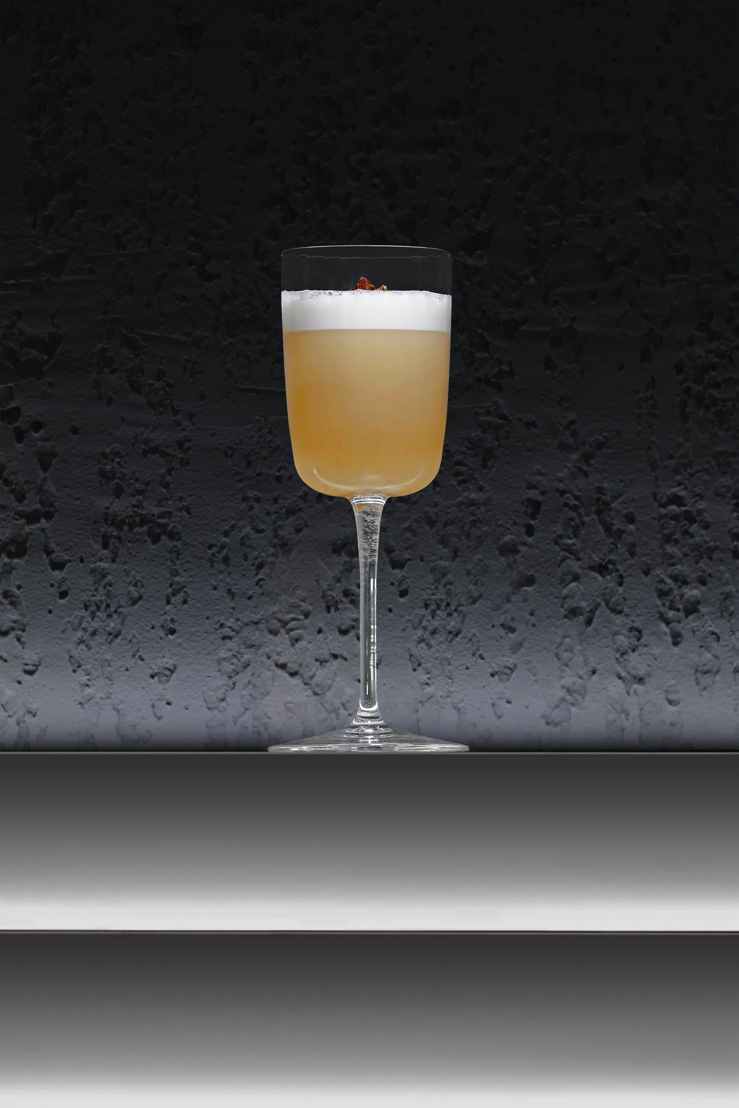 A champagne flute glass with a layered cocktail, topped with foam and garnished with a small red fruit, set against a dark textured background.