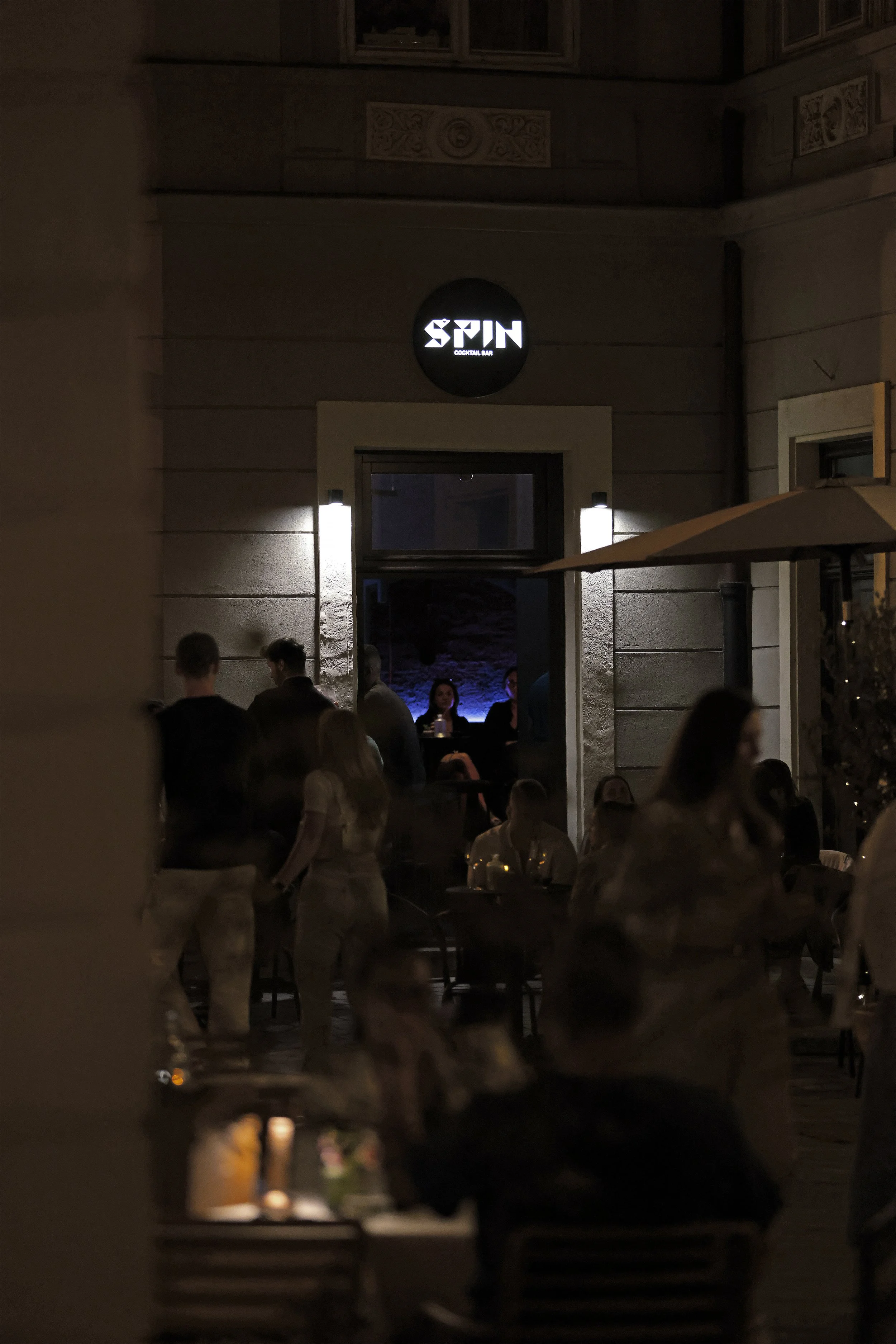 Nighttime scene outside of a bar named 'Spin', with people sitting and standing, some talking and others walking; illuminated signage and ambient lighting create a lively atmosphere.
