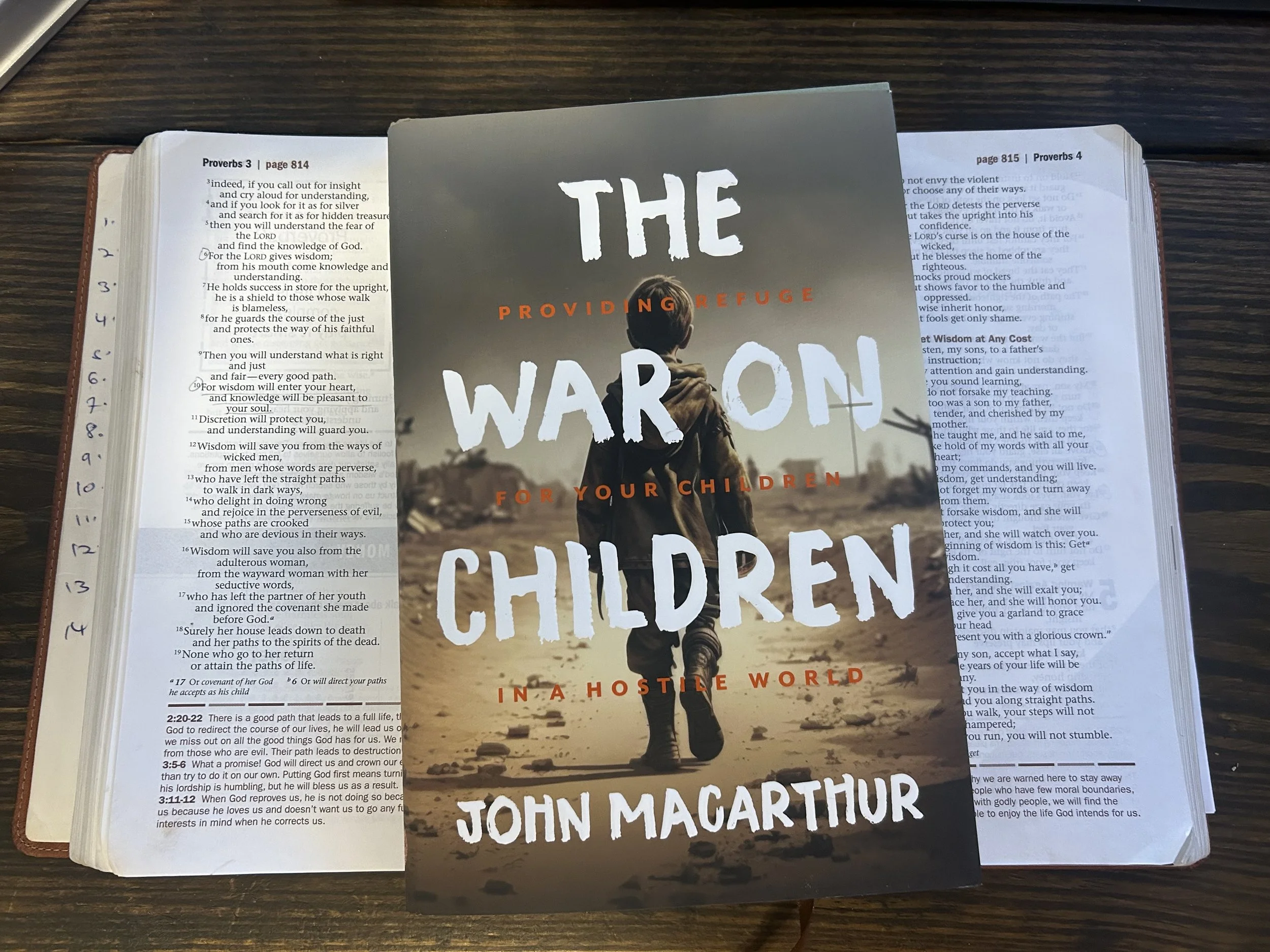 Book titled "The War on Children in a Hostile World" by John MacArthur placed on an open Bible on a wooden surface.