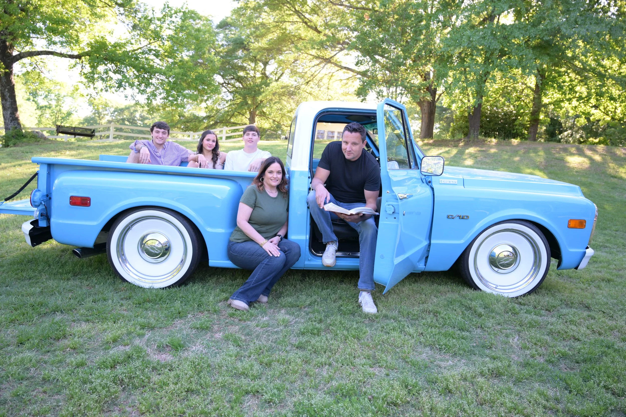 A group of five people outdoors around a vintage blue pickup truck on a grassy area with trees in the background.