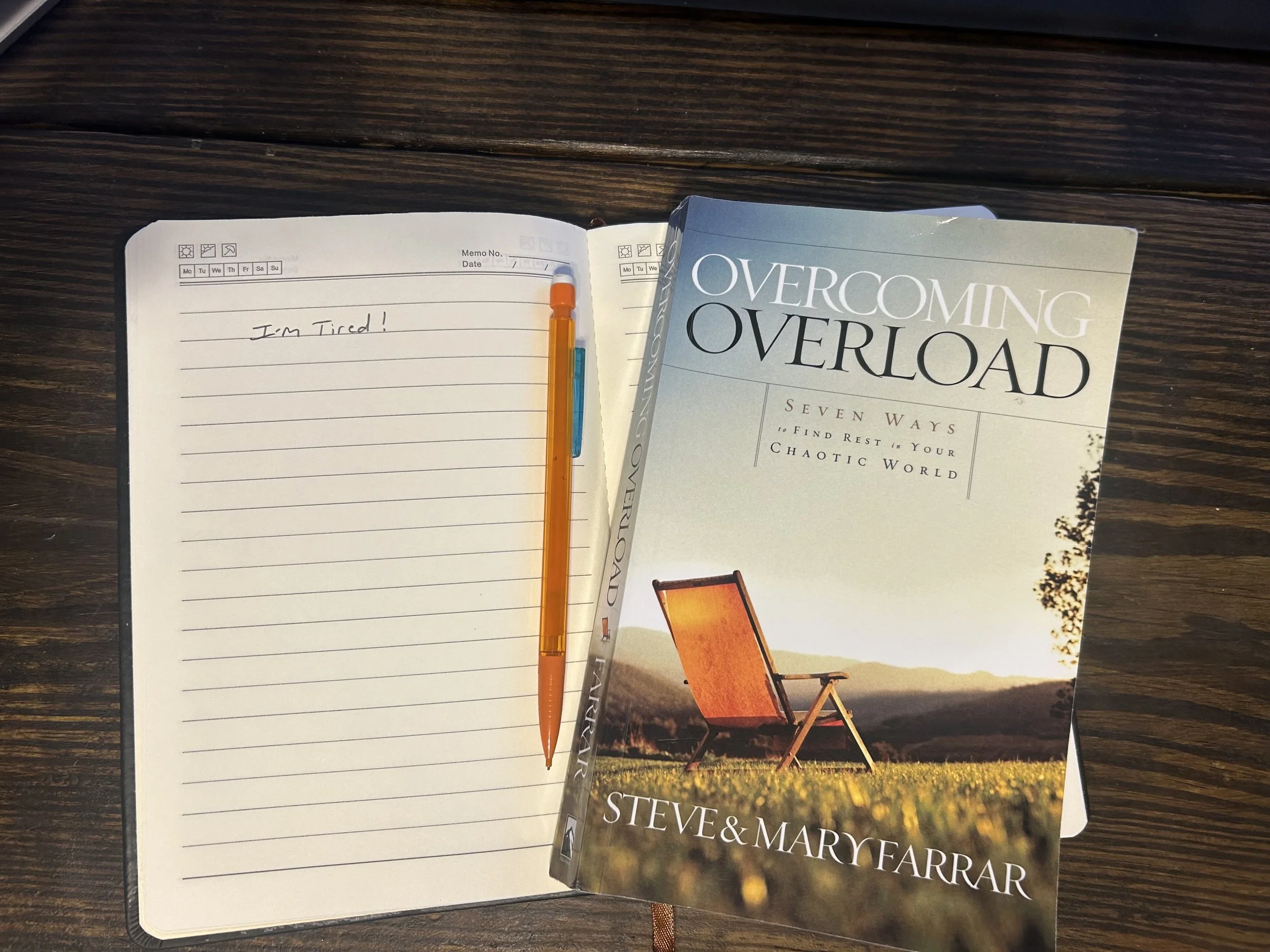 Open planner with handwritten note reading 'I'm Tired!' and four colored pens resting inside. Next to the planner is a book titled 'Overcoming Overload' by Steve and Mary Farrar featuring a landscape with a chair on grass during sunset.