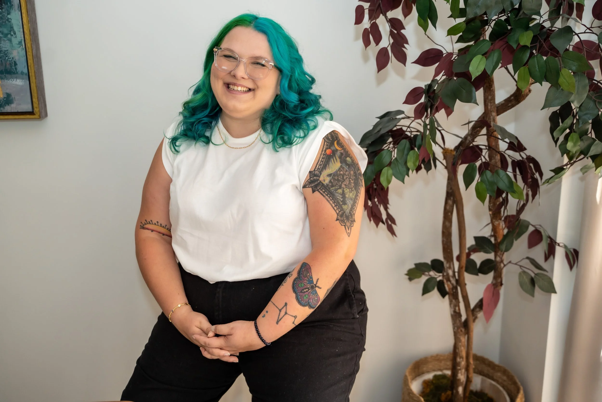 Person with turquoise hair and tattoos smiling, wearing a white shirt and black pants, next to a potted plant.