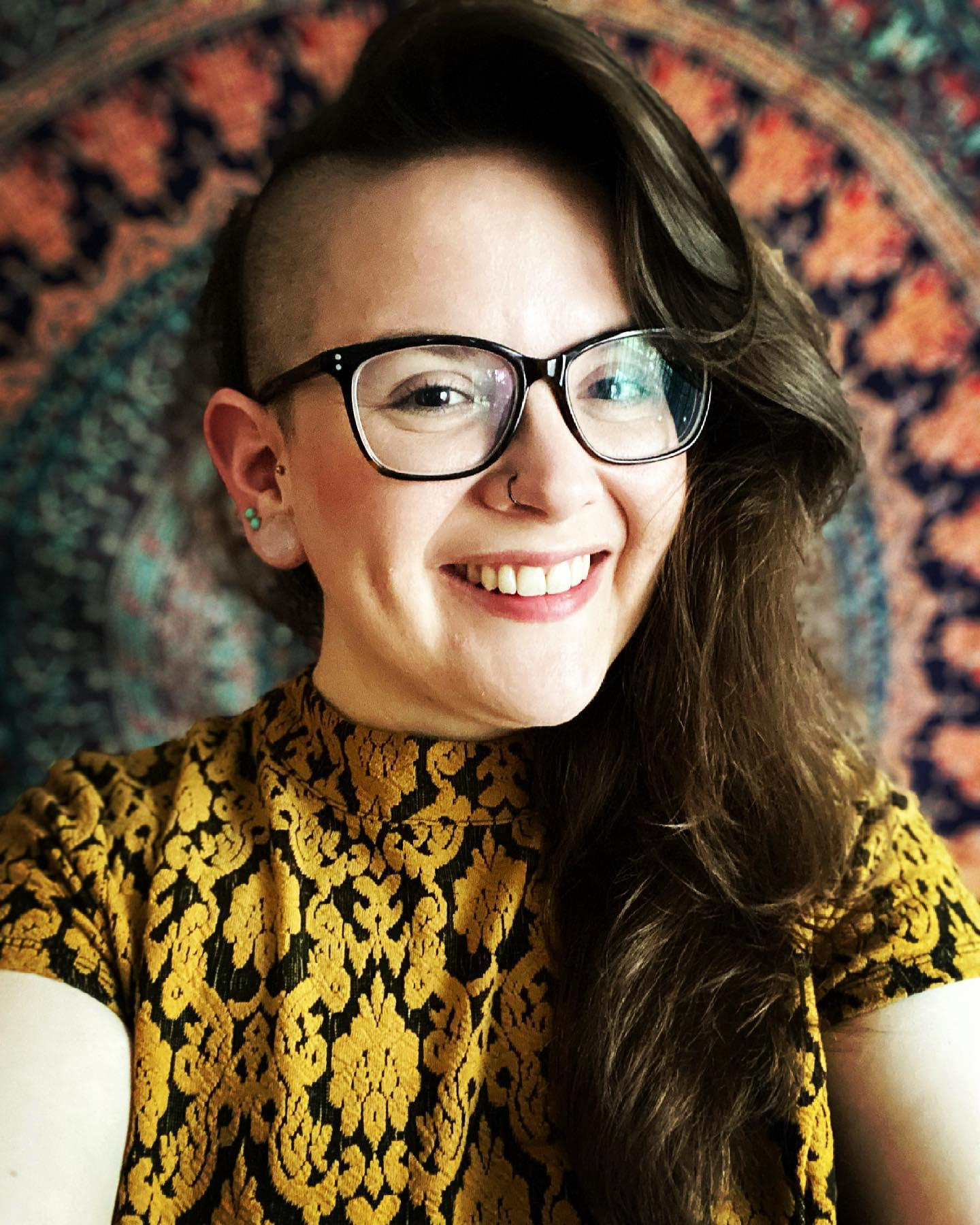 A woman with long brown hair, styled with an undercut on one side, wearing black glasses, a gold and black patterned top, and smiling, with a colorful tapestry or rug in the background.